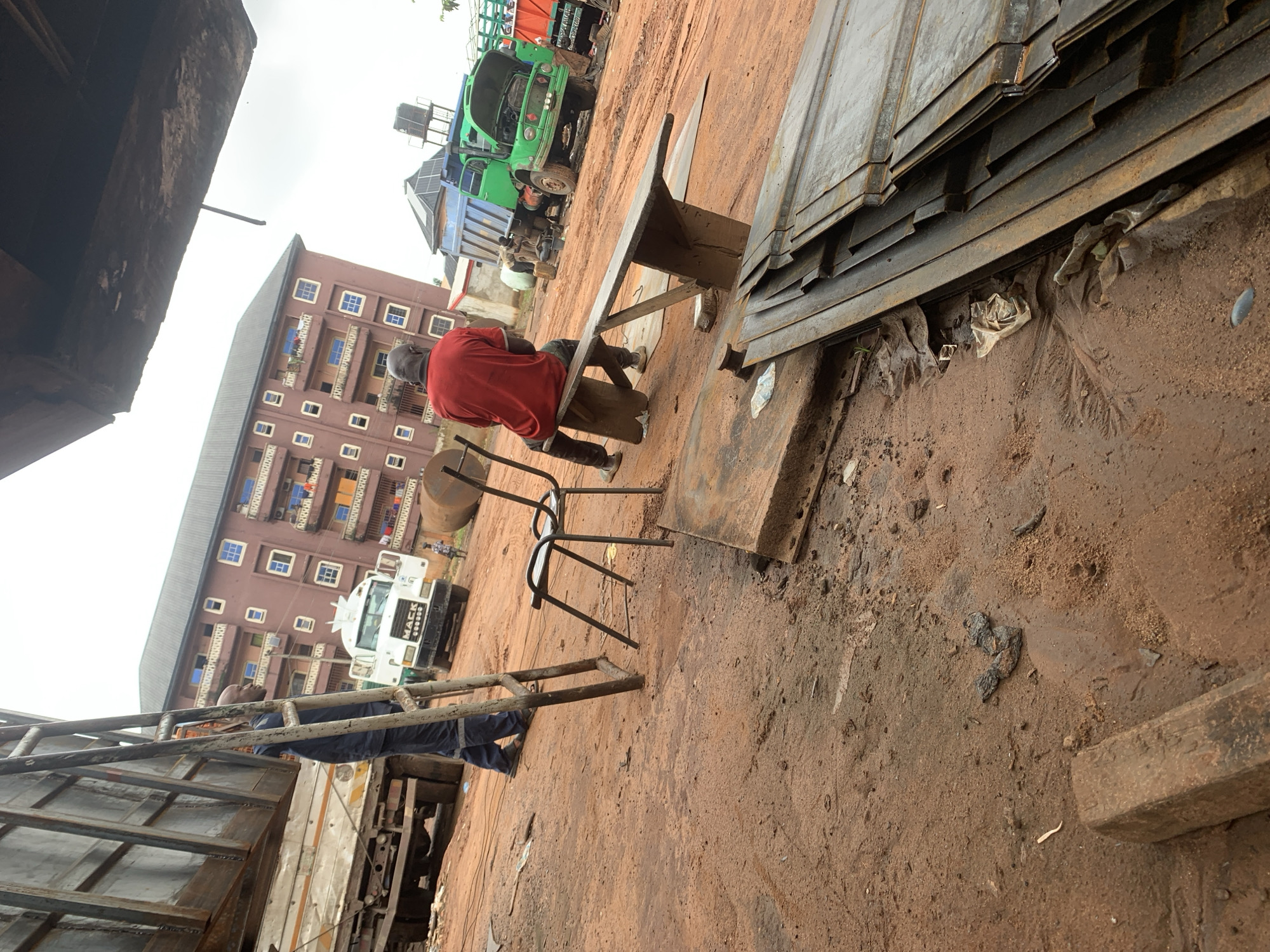 The image depicts an outdoor scene in the city of Nkpor, Nigeria, likely a workshop or storage area for materials. In the foreground, a stack of flat, metallic sheets is piled on the ground, with some crumpled plastic bags scattered nearby. The ground itself is reddish-brown, appearing to be a mixture of soil and dirt. To the left, a person in blue work attire stands near a large, metallic truck. In the middle ground, a man in a red shirt is seated on a makeshift bench, facing away from the camera. Behind him, a metal chair is visible. Further in the background, a white truck with "MACK" branding is parked, and a small child can be seen walking. To the right, a green dump truck with its hood open is parked. A multi-story building with numerous windows and balconies forms the backdrop. The sky is overcast. The overall impression is a busy, working environment with industrial elements.