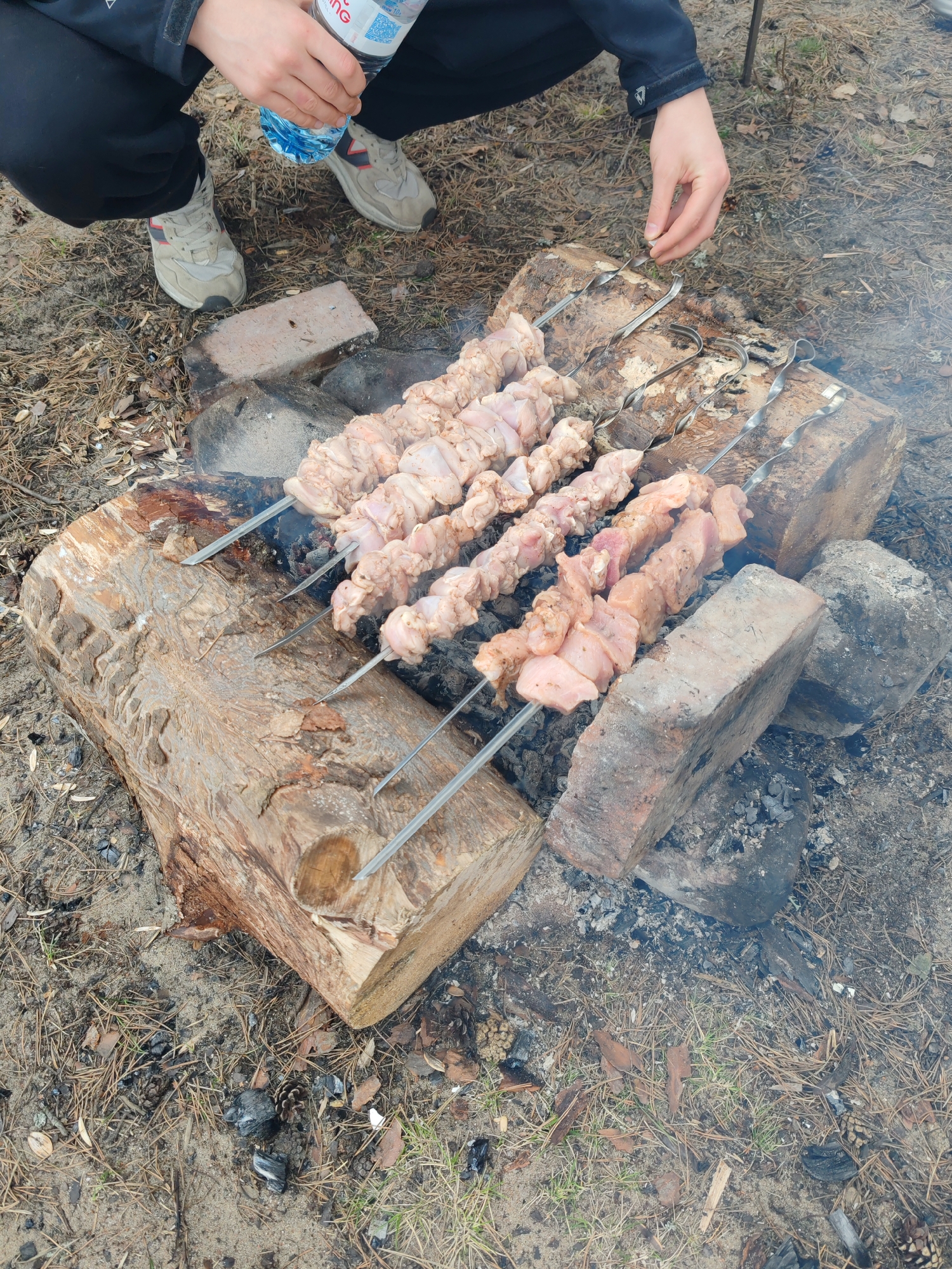 In an outdoor setting near Bolshaya Izhora, Russia, a person wearing black pants, grey sneakers, and a dark blue jacket crouches near a makeshift barbecue. They hold a plastic water bottle in their left hand while their right hand rests above metal skewers laden with raw meat, positioned over a fire built on a large, roughly hewn log supported by red bricks and charred remnants. The ground is sandy and littered with pine needles and twigs. Smoke rises from the smoldering fire.