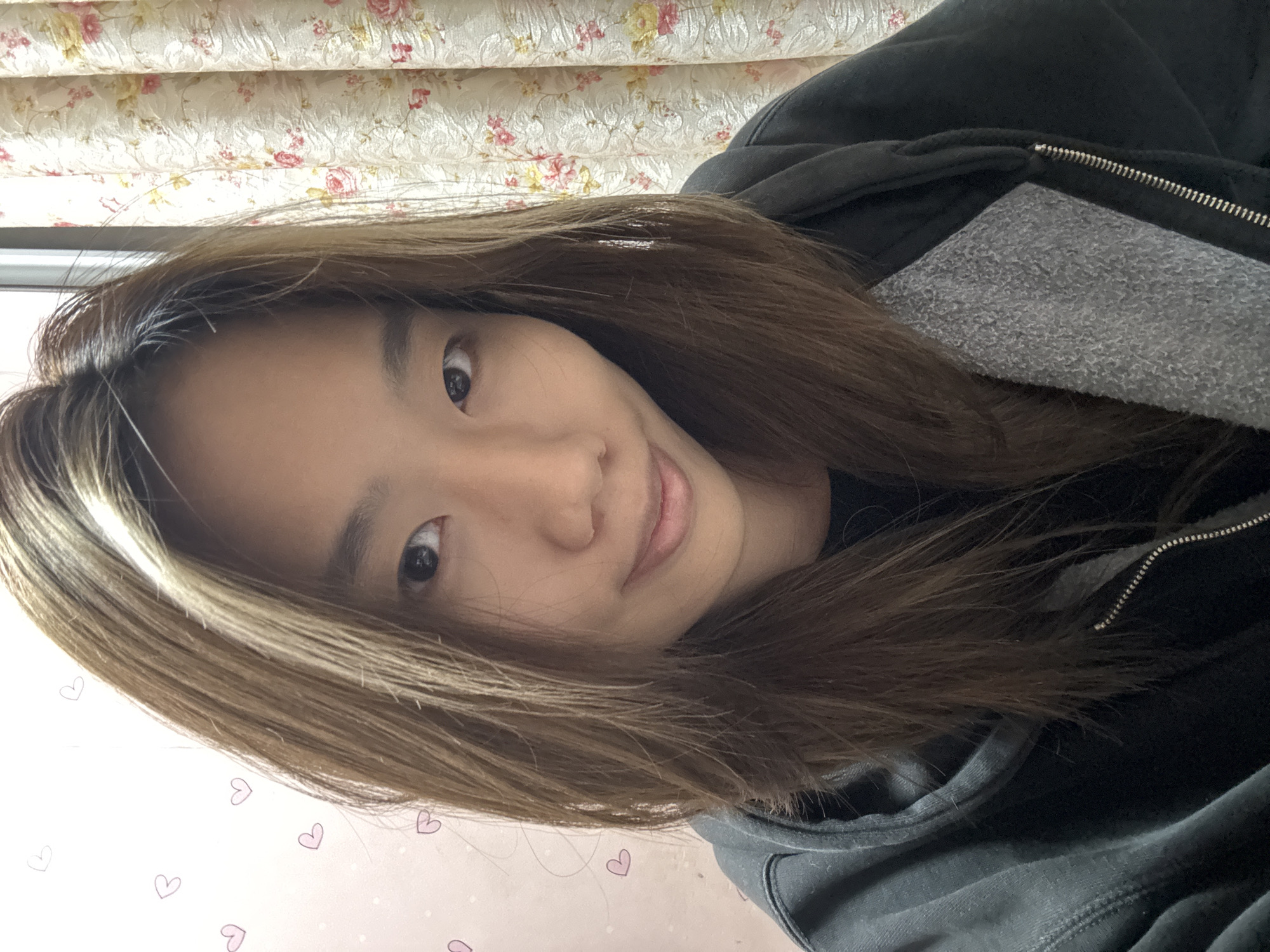 A selfie of an Asian woman with long brown hair is presented at an angle. The woman is wearing a dark hooded sweatshirt with a visible gray lining. Her head is tilted slightly, and she looks directly at the camera with a soft smile. In the background, there is a floral patterned curtain and a light pink wall with small heart designs. While there's nothing to directly indicate the location as Shapingba District, China, it can't be ruled out.