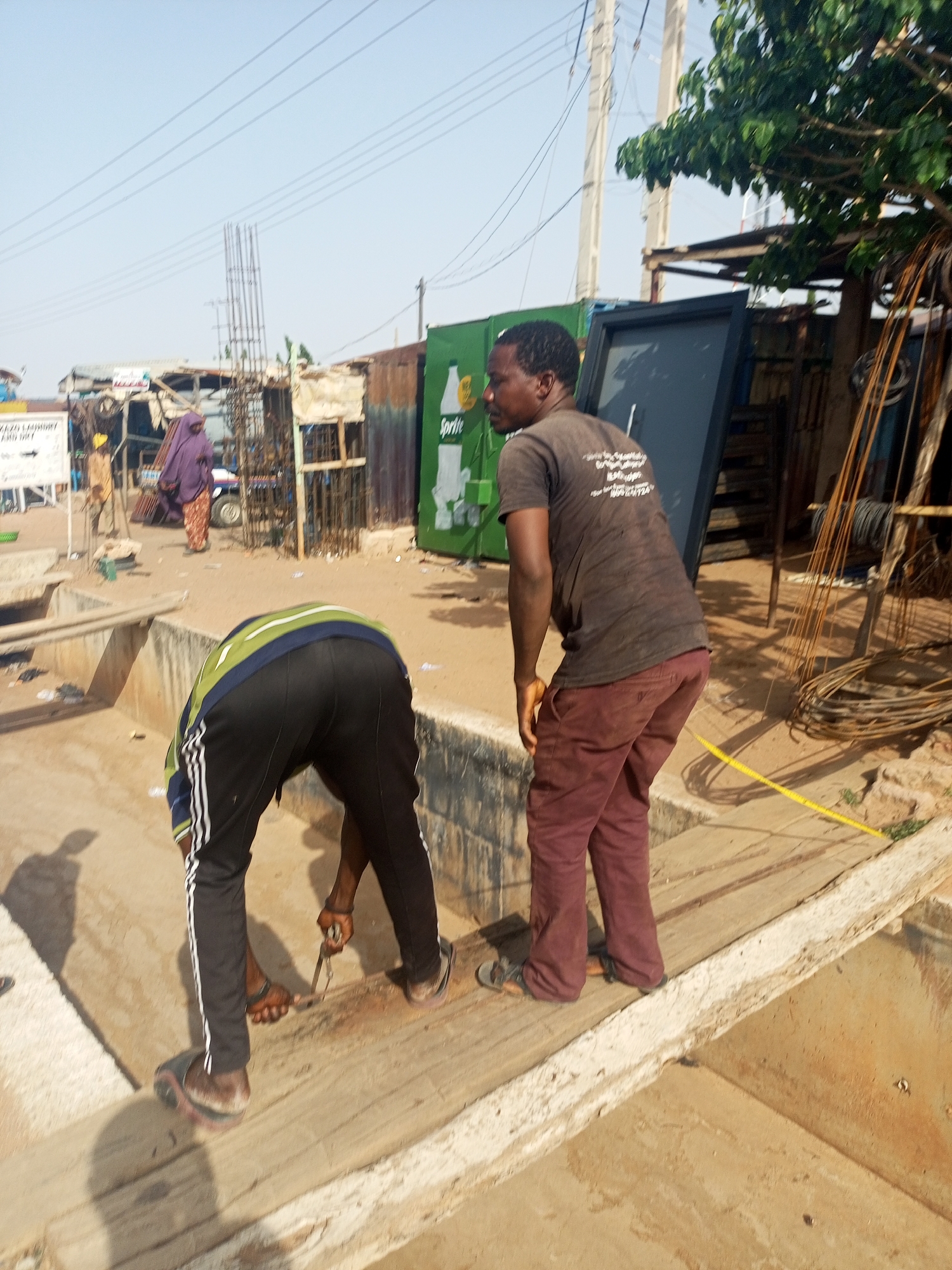 Two men stand near a concrete structure. One man bends over, manipulating something with a tool. The other man stands upright, looking to the side. In the background, there is a street scene with various buildings, including a structure with exposed rebar. A woman wearing a purple outfit walks along the street. A sign is visible. The environment appears to be an urban area in Tempure, Nigeria. Steel rods are present behind the standing man. A yellow measuring tape is stretched across the concrete structure.