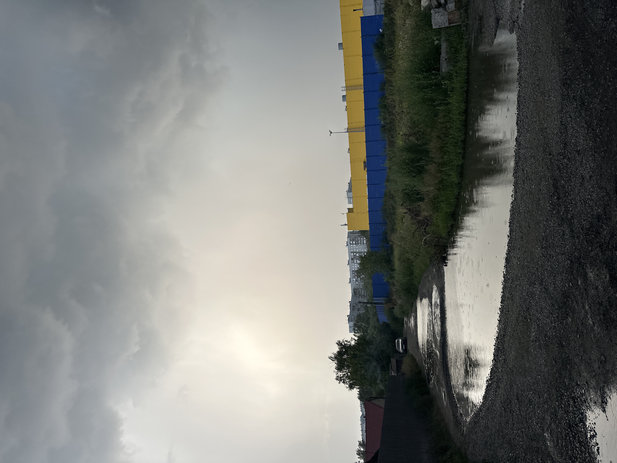 The image depicts an overcast sky with the sun breaking through the clouds on the left side, creating a bright spot. In the foreground, a dirt road with numerous puddles, indicative of recent rain, stretches into the distance. To the left of the road, a dark metal fence runs alongside residential buildings with red roofs. Further down the road, a dark-colored car is visible. On the right side of the road, there is dense green vegetation bordering a long, colorful fence, which is divided into sections of bright yellow and blue. Beyond the fence, there are multi-story apartment buildings in the background, suggesting an urban or suburban environment. The scene is likely located in Biysk, Russia, given the typical architecture and weather patterns.