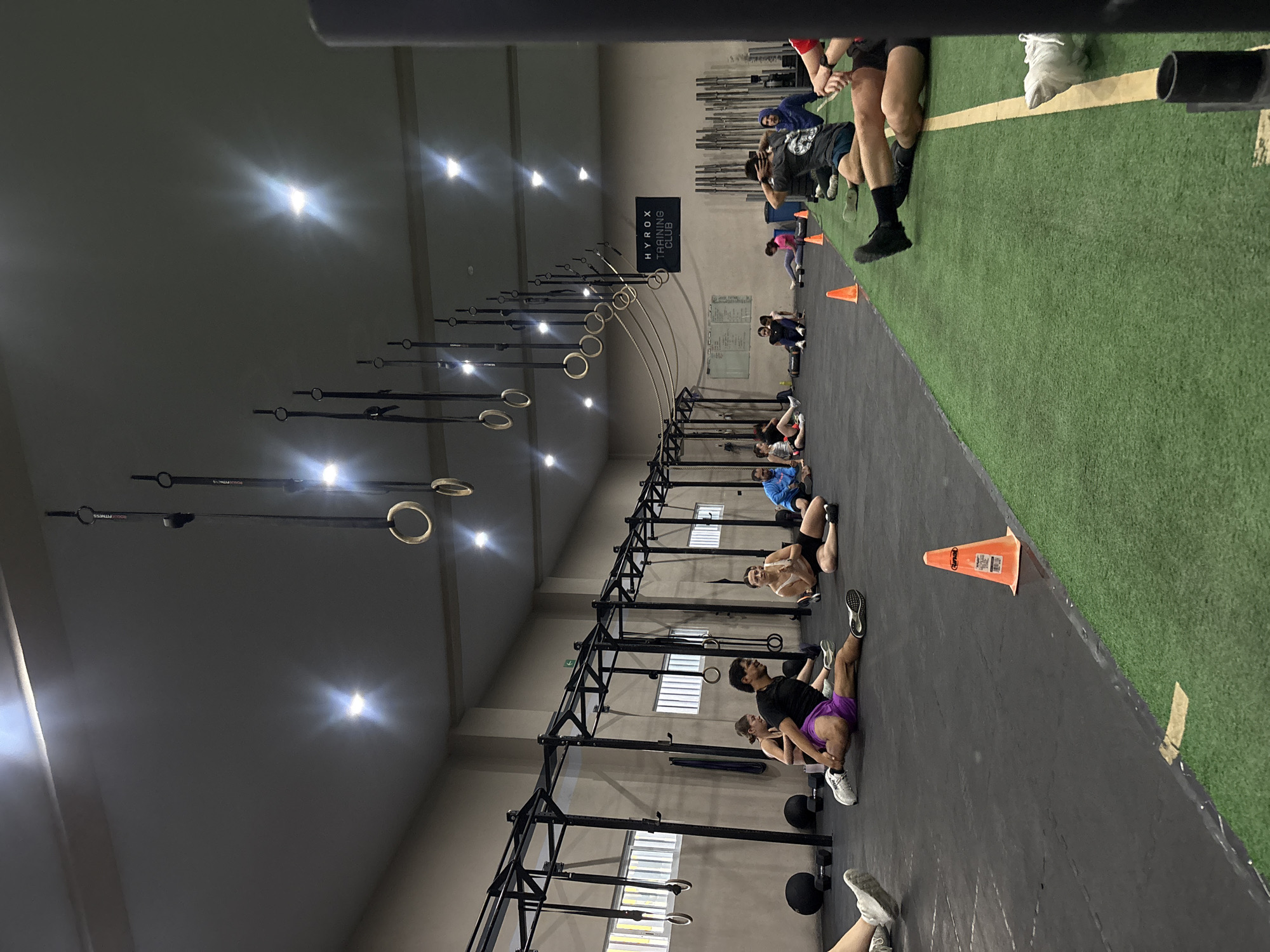 The image shows an indoor gym setting, potentially located in San Bernardino Tlaxcalancingo, Mexico (location not definitively confirmed within the image itself). Several people are engaged in physical exercises on a black floor area. The gym includes athletic equipment such as pull-up rigs, hanging rings, and an adjacent green artificial turf area. Cones are visible marking the workout zones. A sign for "HYROX TRAINING CLUB" hangs in the background. The environment is well-lit with ceiling lights.