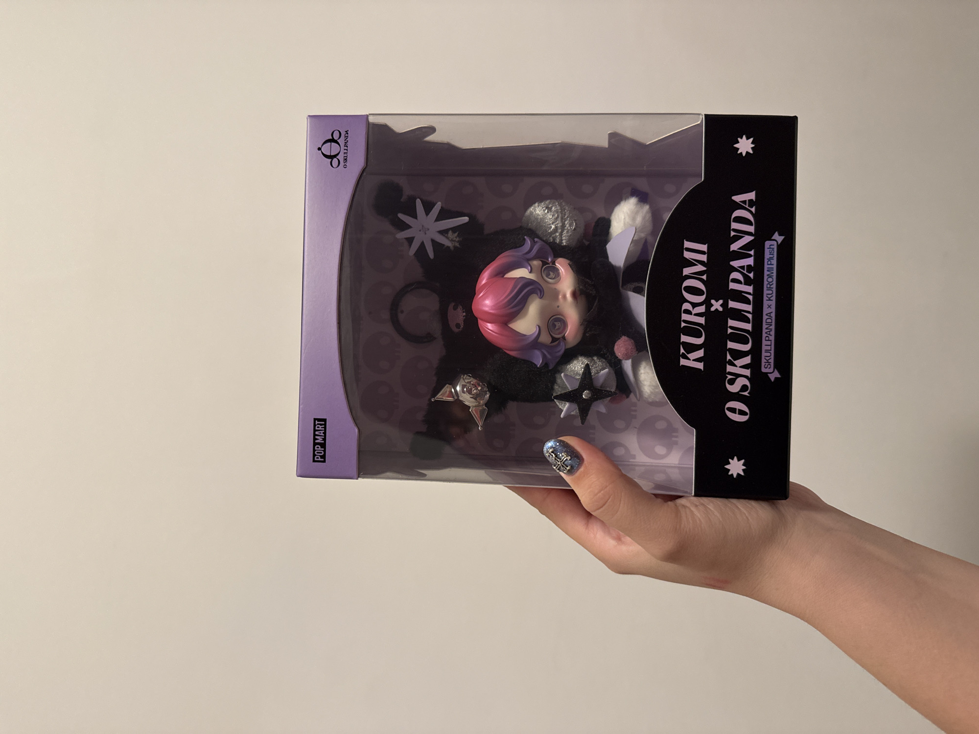A person is holding a packaged Skullpanda x Kuromi plush toy against a light beige background. The toy is in a clear plastic and black box. The front of the box reads "KUROMI X O SKULLPANDA" and includes the text "SKULLPANDA x KUROMI Plush." The side of the box reads "POP MART" and "O SKULLPANDA". The person's hand and part of their arm are visible. Their fingernails are decorated with blue glitter and a silver cross design.
