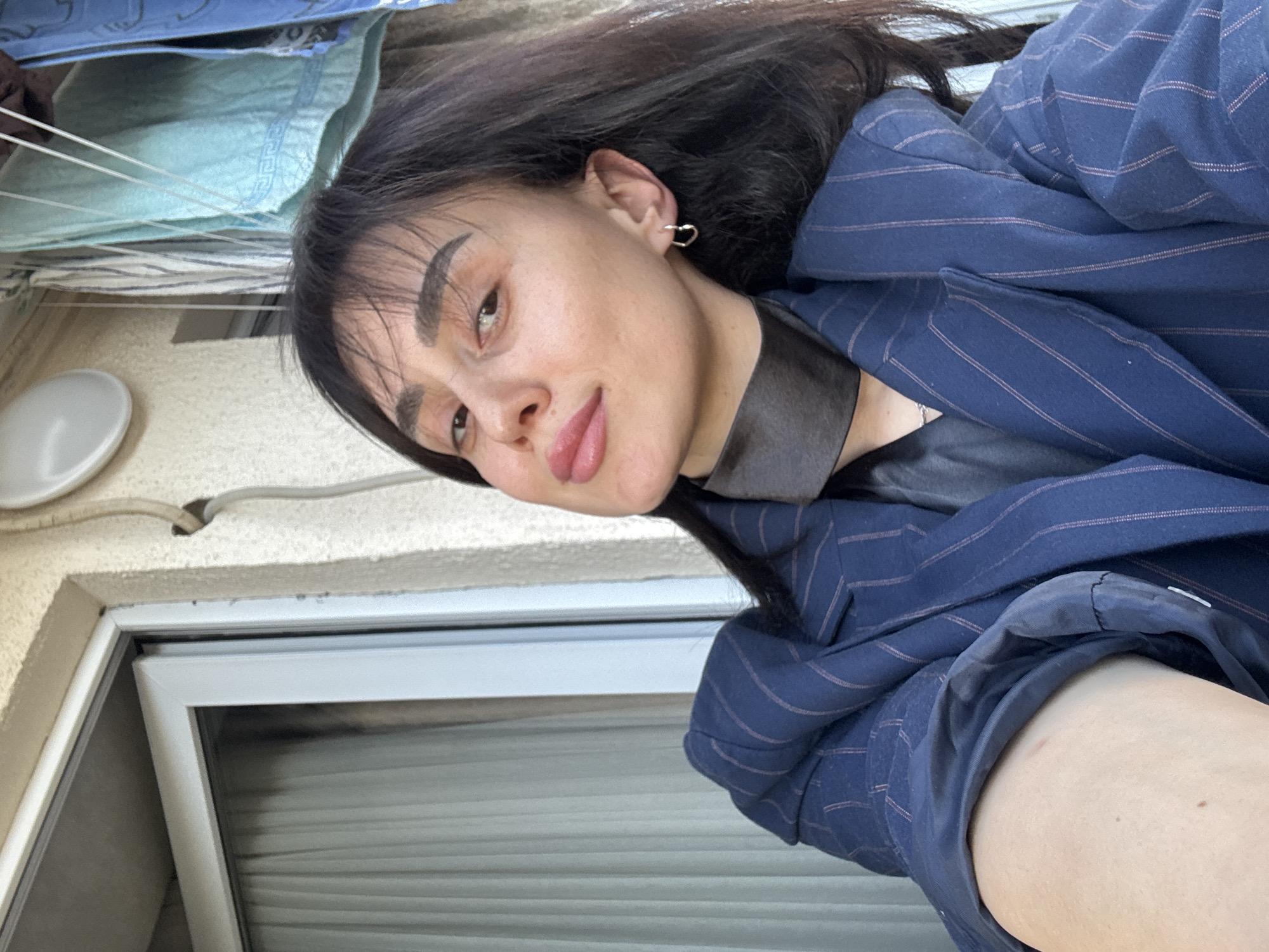 A woman is pictured in a close-up, angled selfie. She has dark hair and is wearing a blue pinstripe blazer over a dark top. She is wearing small, heart-shaped earrings. The background shows a white wall, a closed window with horizontal blinds, and a clothesline with some hanging fabrics. The location is Tbilisi, Georgia, based on the architectural style and the presence of a clothesline hanging outside a window.