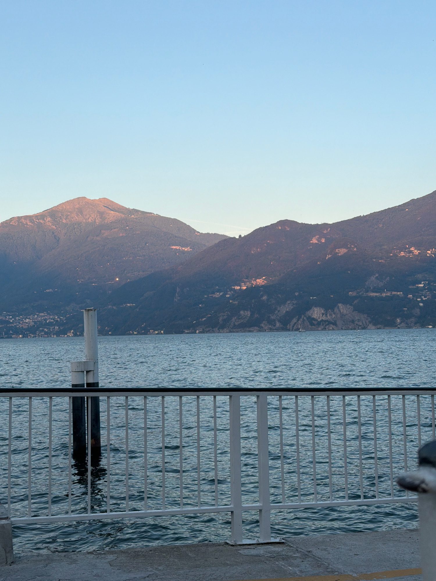 The image depicts a scenic view of a lake with mountains in the background, likely taken from a waterfront area in Loveno, Italy. The foreground features a white metal railing with vertical bars, separated by a concrete walkway. To the left, a white post with black banding stands in the water. The water itself is calm with gentle ripples. Beyond the railing and water, a village is visible nestled on the slopes of the dark, tree-covered mountains. The peaks of these mountains are illuminated by the warm light of either sunrise or sunset, casting a soft glow on their upper portions. The sky is a clear, pale blue. The overall impression is one of tranquility and natural beauty.