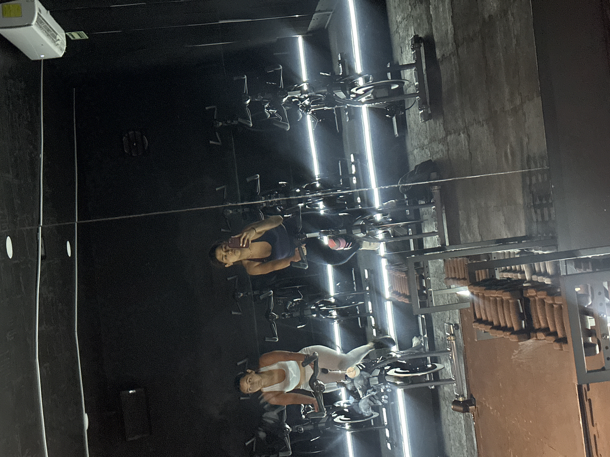 A photograph taken in San Bernardino Tlaxcalancingo, Mexico, shows two women riding stationary bikes in a gym setting. One woman wears a white tank top and light-colored leggings, and the other wears a dark tank top and dark leggings. The gym is lit by bright, vertical light bars. The image appears to be a reflection, as there are multiple rows of stationary bikes and other gym equipment visible in the background. One of the women is holding a mobile phone in her hand, suggesting she may be taking a photograph of herself. The gym walls are dark in color.
