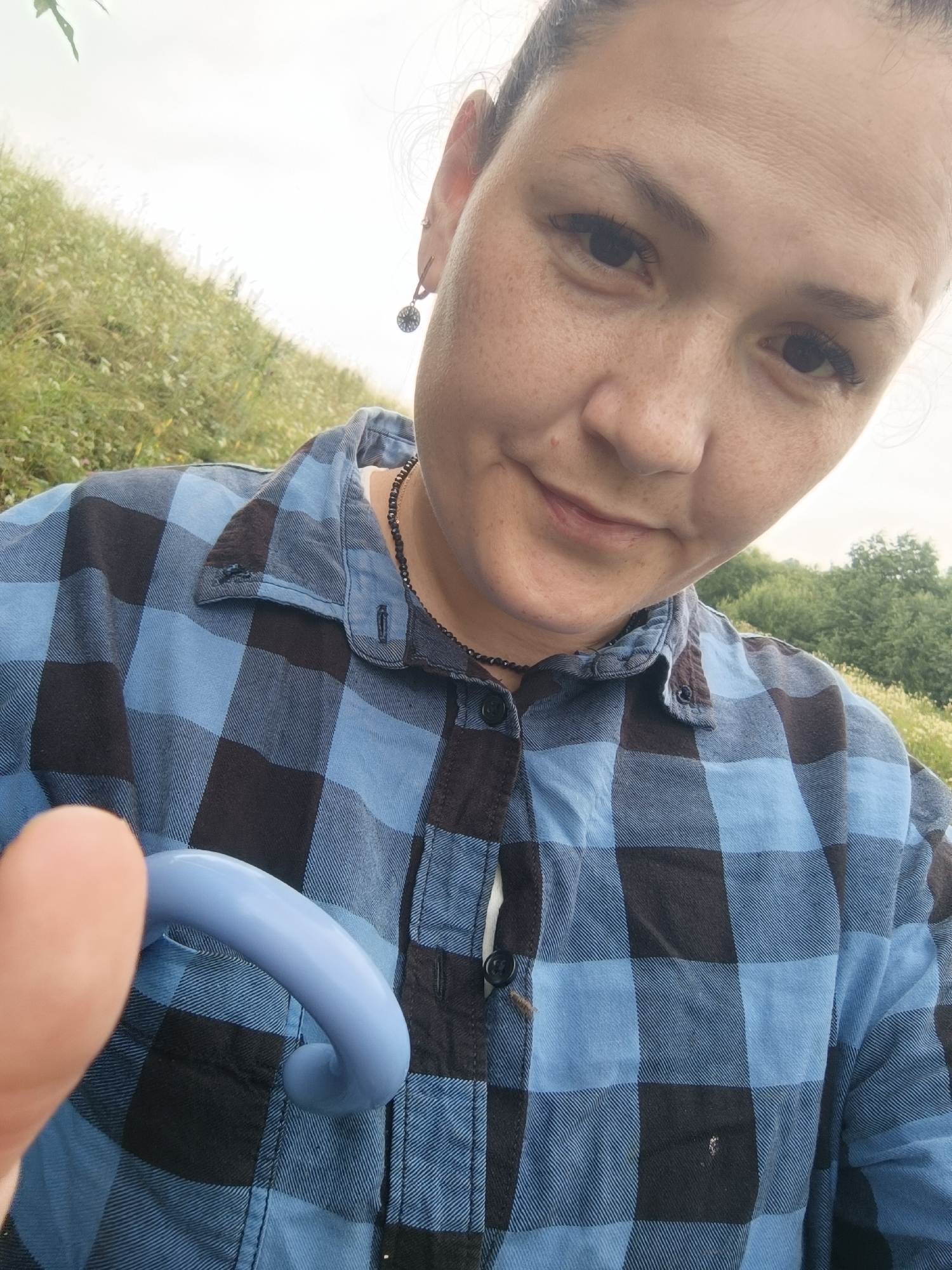 A woman is taking a selfie outdoors on a cloudy day. She is wearing a blue and black plaid shirt and a black beaded necklace. Her left ear has a small, round earring. In the foreground, her left thumb is partially visible, obscuring a light blue, curved object. The background is a grassy hillside with some trees in the distance. This scene is not located in the city of Karsha, Russia.