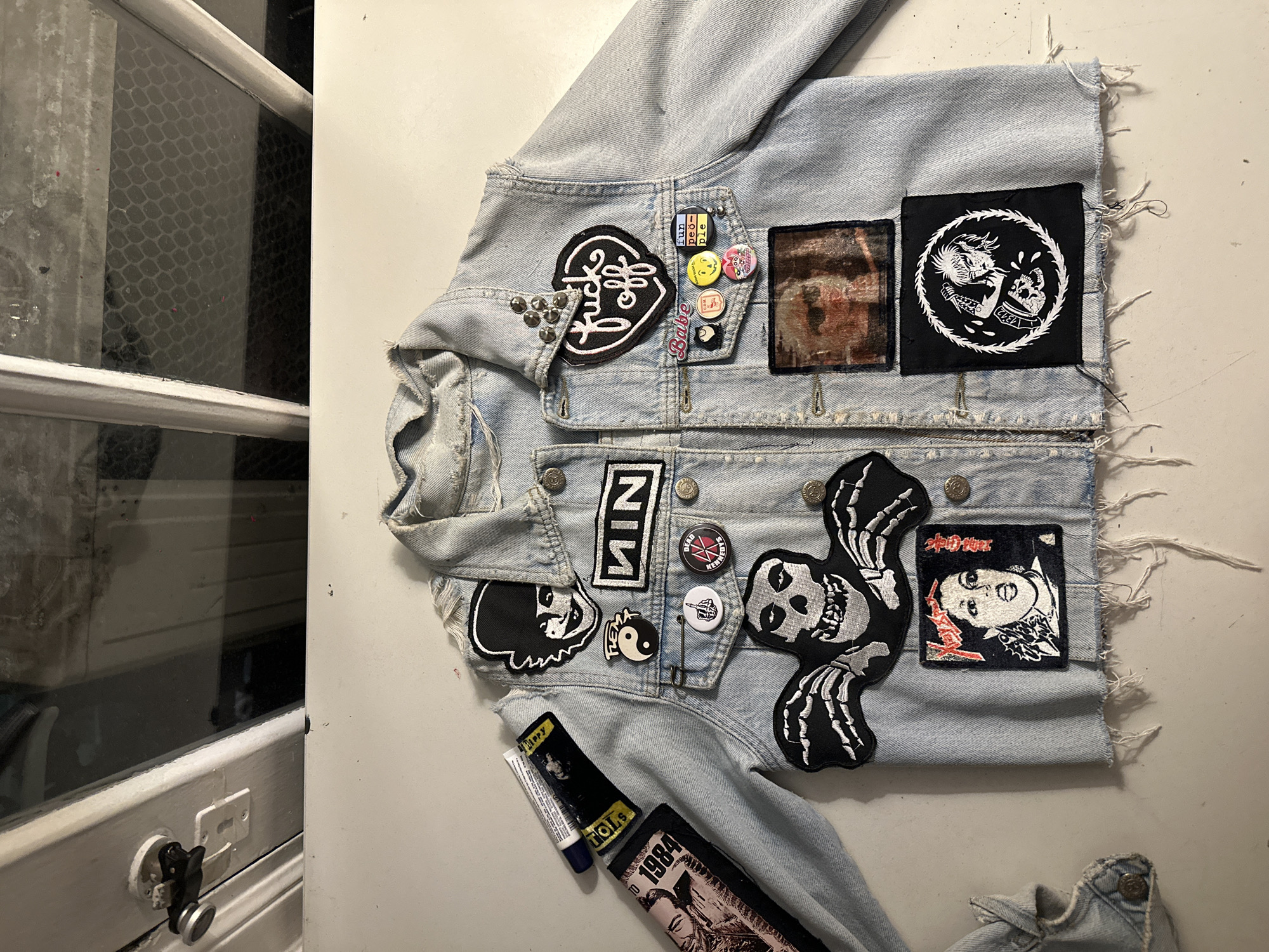 A light blue denim jacket with patches and pins lies on a white surface near a window. The jacket has various patches, including band patches, a skull with hands, and political symbols. Several pins are clustered around the top right chest area. Below the jacket's left sleeve, a tube of glue and a patch are visible. The window has a grid pattern and is partially obscured by a white frame and wall. The scene appears to be indoors.