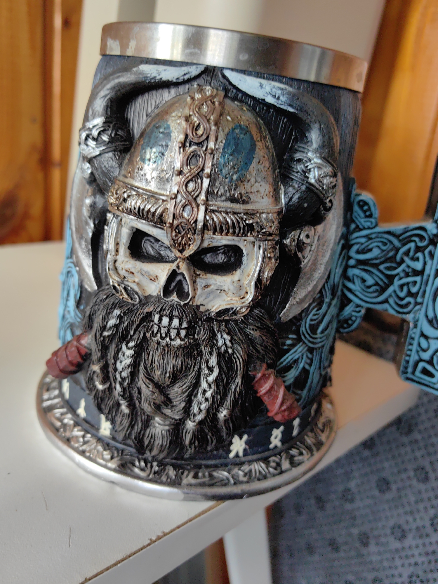 A detailed image depicts a heavily ornamented decorative tankard or mug, made from dark, textured resin or similar material, featuring a silver-toned metal rim at the top and base. The central design on the mug's body is a detailed skull wearing an aged, metallic Viking-style helmet. The helmet is primarily silver-colored with bronze accents and exhibits patches of teal-blue patina, indicative of weathering or intentional aging. Intricate knotwork patterns are etched into the helmet's brow and sides. The skull itself is off-white or bone-colored, with deep black eye sockets and a visible skeletal nasal cavity. A long, dark, braided beard with lighter highlights extends from the skull's jawline, featuring red-wrapped elements on its lower left side. Surrounding the central skull are dark, sculpted elements, possibly depicting intertwined serpents or additional knotwork. The handle of the mug, situated on the right, is shaped like an axe, with a silver-colored blade and a striking turquoise-blue handle adorned with elaborate Celtic or Viking scrollwork. The mug rests on a flat, clean white surface, likely a tabletop or shelf. The background is out of focus, revealing warm, blurred reddish-brown tones, suggestive of wooden paneling or furniture, on both the left and right sides. A darker, patterned surface is partially visible on the far right, below the level of the mug's handle. The scene depicts an object in an indeterminate indoor setting, located in the city of Unknown, Unknown.