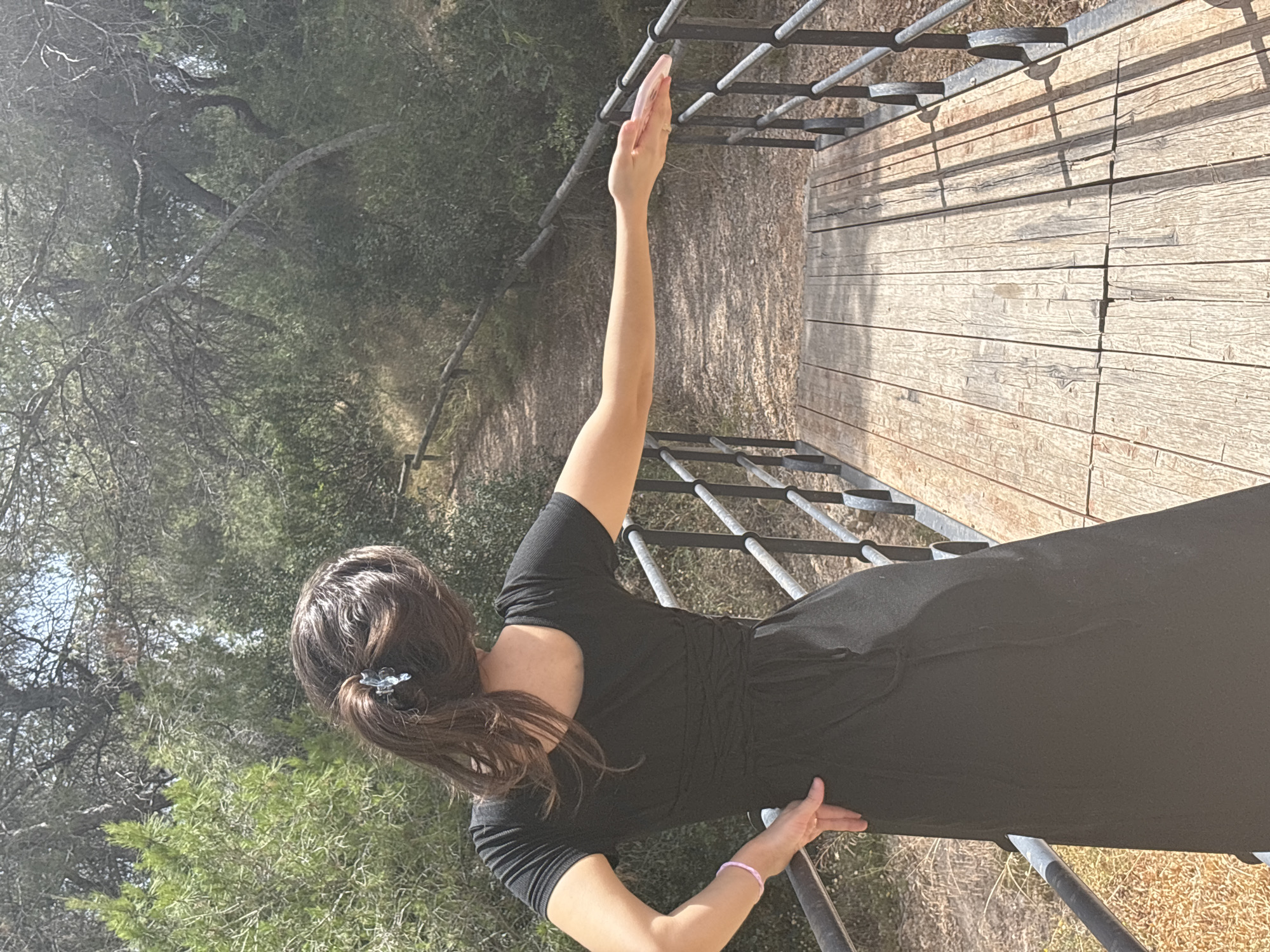 A person is shown from the back, standing on a wooden platform. The person has long, dark brown hair tied in a ponytail with a translucent clip. They are wearing a black dress and a thin pink bracelet on their left wrist. Their right arm is raised upwards and slightly bent, with their hand open and palm facing forward. The person is positioned next to a metal railing with horizontal bars.
The setting appears to be outdoors, with lush green foliage and trees visible in the background. The wooden platform is made of weathered planks, and shadows from the railing are cast upon it. The lighting suggests it is daytime, likely morning or afternoon, with sunlight filtering through the trees. The overall mood is serene and peaceful. There is no visible text in the image.