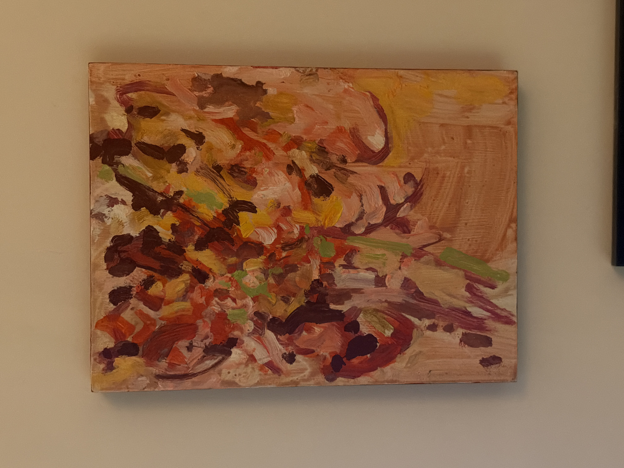 An abstract painting, rendered with prominent, impasto-style brushstrokes in a palette dominated by reds, oranges, yellows, browns, and lighter beige/pink tones with green accents, is centrally positioned on a plain, light beige wall. The painting appears unframed or thinly gallery-wrapped, revealing the edges of the canvas. A subtle shadow is cast by the bottom edge of the artwork onto the wall. A dark, thin, horizontal object is partially visible at the bottom of the overall image, below the painting's frame. The scene is located in the city of London, United Kingdom.