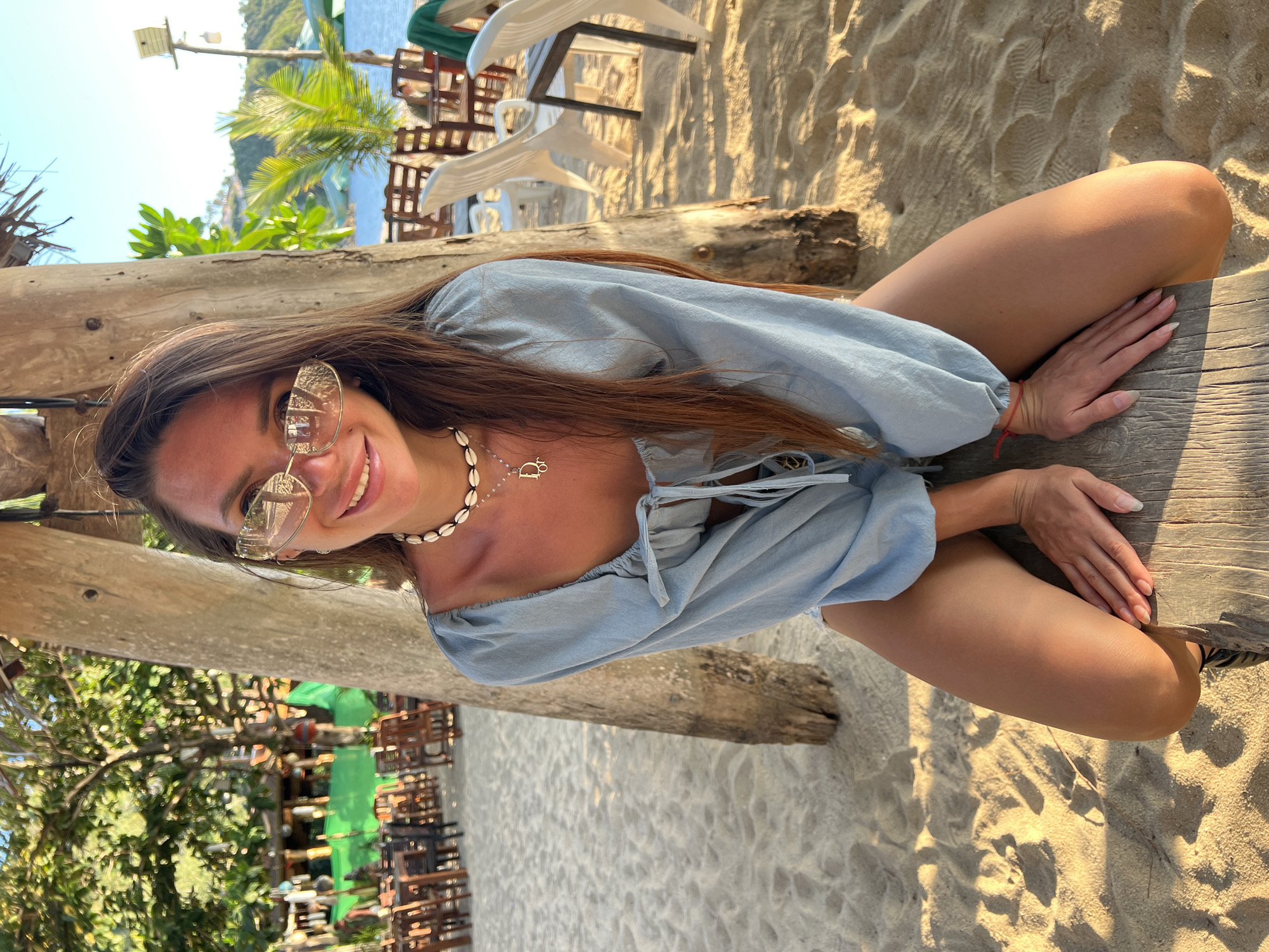 A woman with long, brown hair and sunglasses sits on a wooden beam on a sandy beach. She is wearing a light blue off-the-shoulder top and appears to be in a relaxed pose. Behind her, palm trees and a body of water are visible. In the background, there are tables and chairs set up, suggesting a beachfront restaurant or bar. The scene is bathed in warm, golden light, indicating it is likely late afternoon or early morning. The location is described as Tambon Ko Phangan, Thailand.