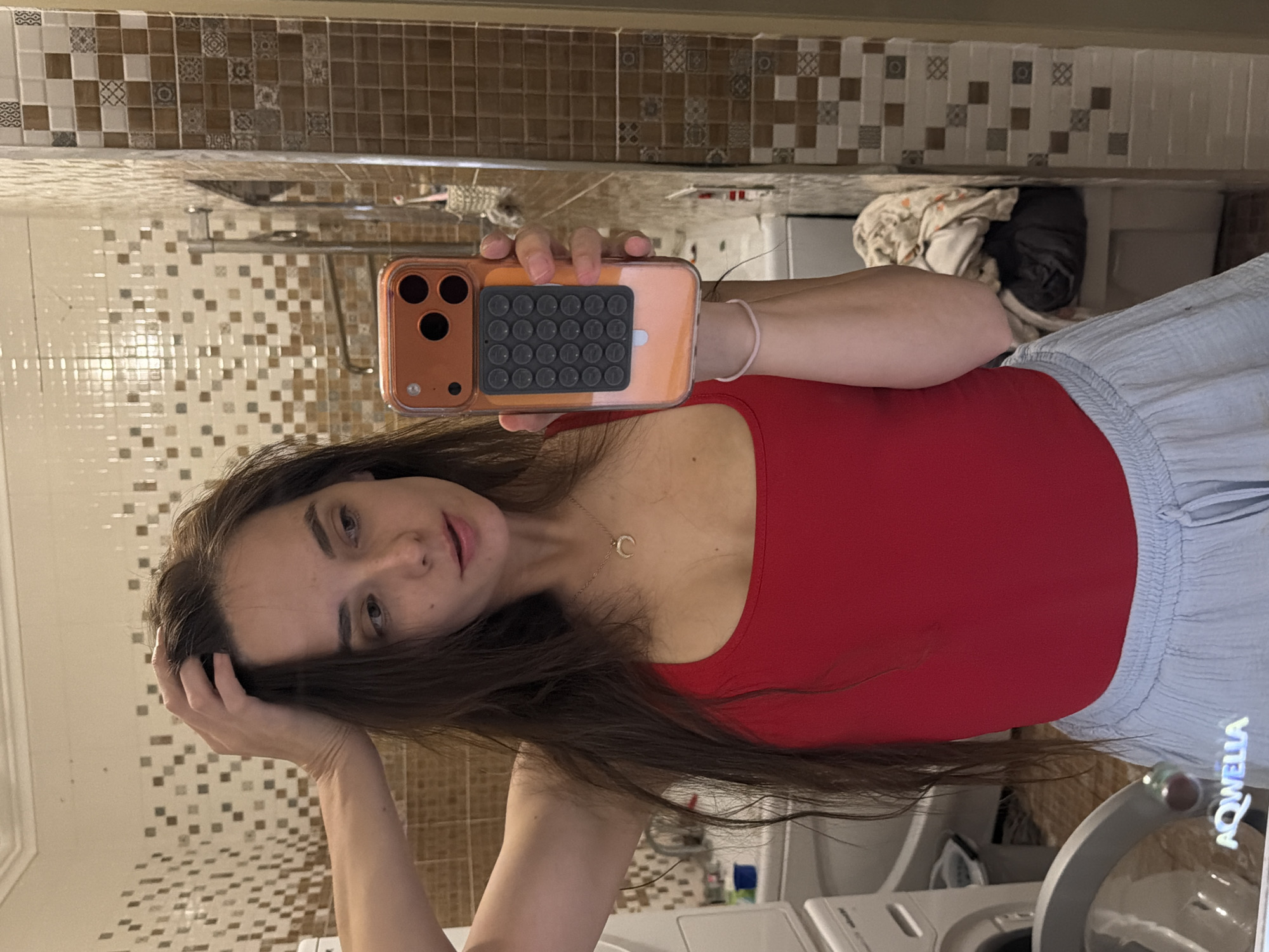 A young woman with long, dark brown hair is taking a selfie in a bathroom. She is wearing a red tank top and a light blue skirt. She has her left hand on her head and is holding an orange phone with her right hand. The phone has a case with a circular grid pattern on the back. The bathroom has tiled walls and a washing machine is visible in the background.