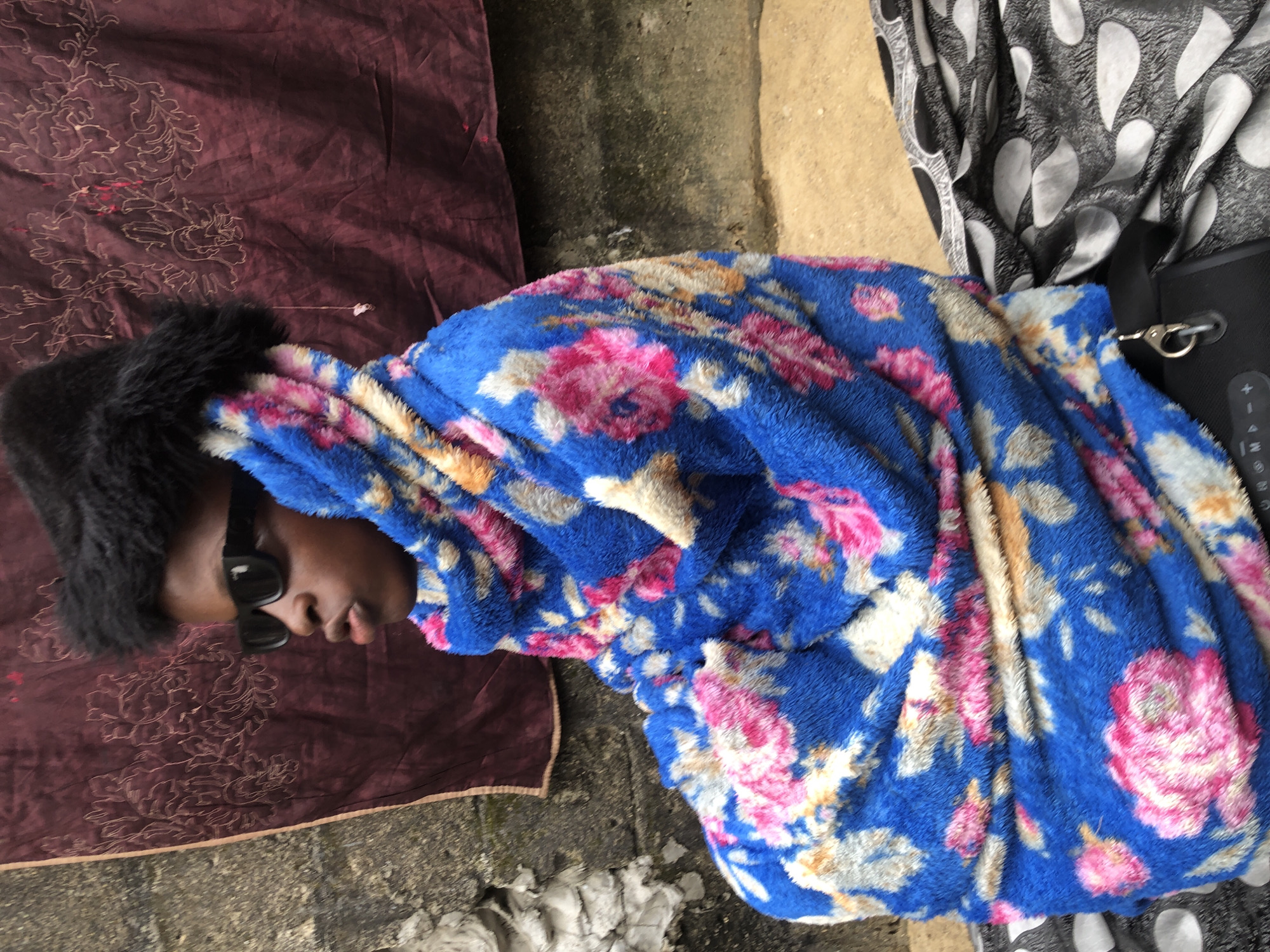 A young person wearing sunglasses and a black furry hat is wrapped in a blue blanket with pink and white floral patterns. They are seated outside against a textured wall, with a dark, patterned fabric draped over their lap. The scene appears to be in Ado, Nigeria, possibly in an outdoor urban setting given the wall textures and exposed ground.