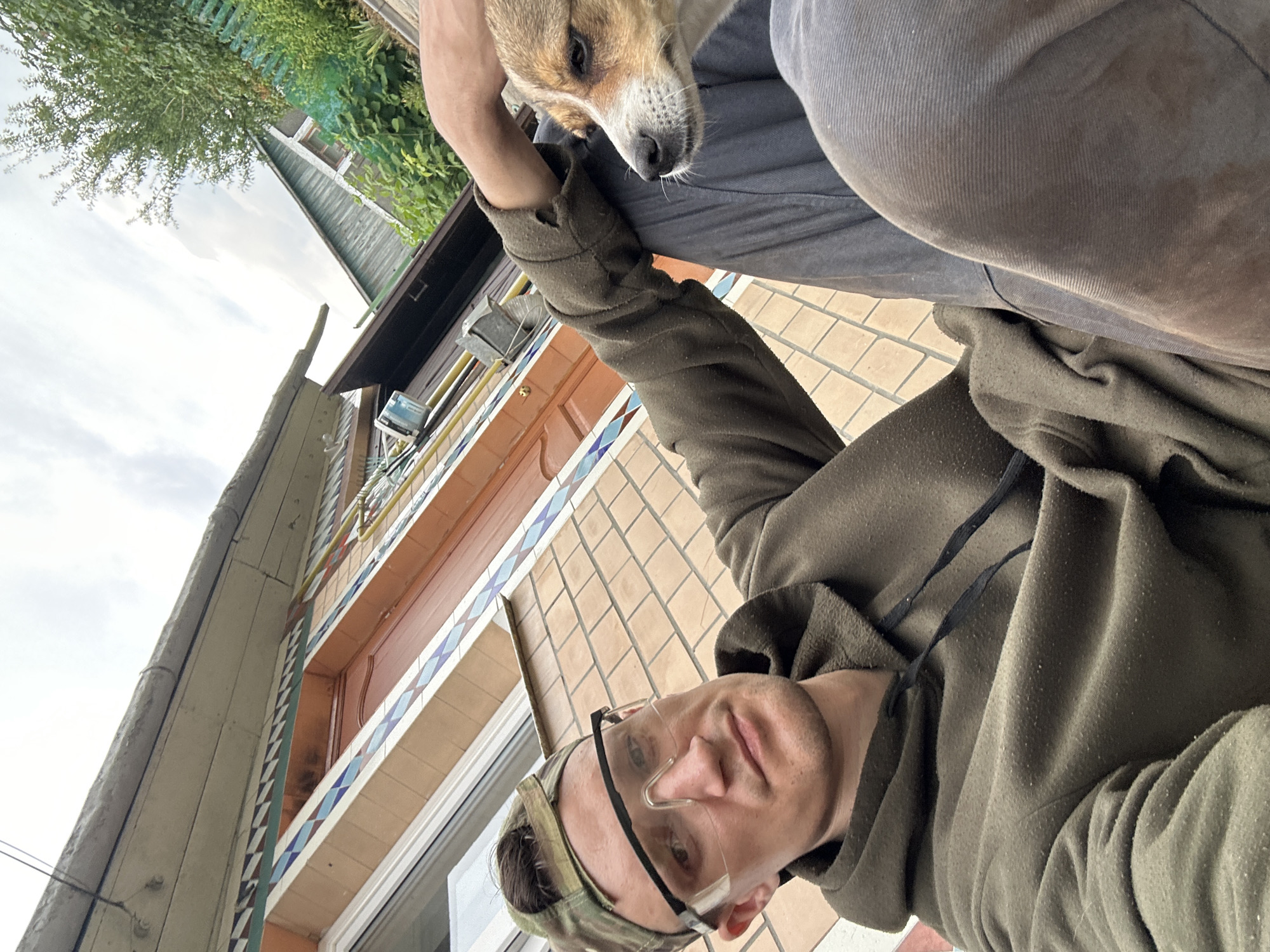 A man wearing a camouflage cap and protective goggles, along with a dark green hoodie, is seated outdoors with a small dog on his lap. He is looking towards the camera, and his left hand is resting on the dog's head. The dog is facing to the right, with its head turned slightly towards the man. The man is wearing dark pants. The background shows a building with tiled walls and a wooden roof, along with some greenery and a cloudy sky. The overall impression is that this is a candid, informal photograph taken in a rural or suburban setting, possibly during a break from outdoor work. The location is identified as Makiv, Ukraine.