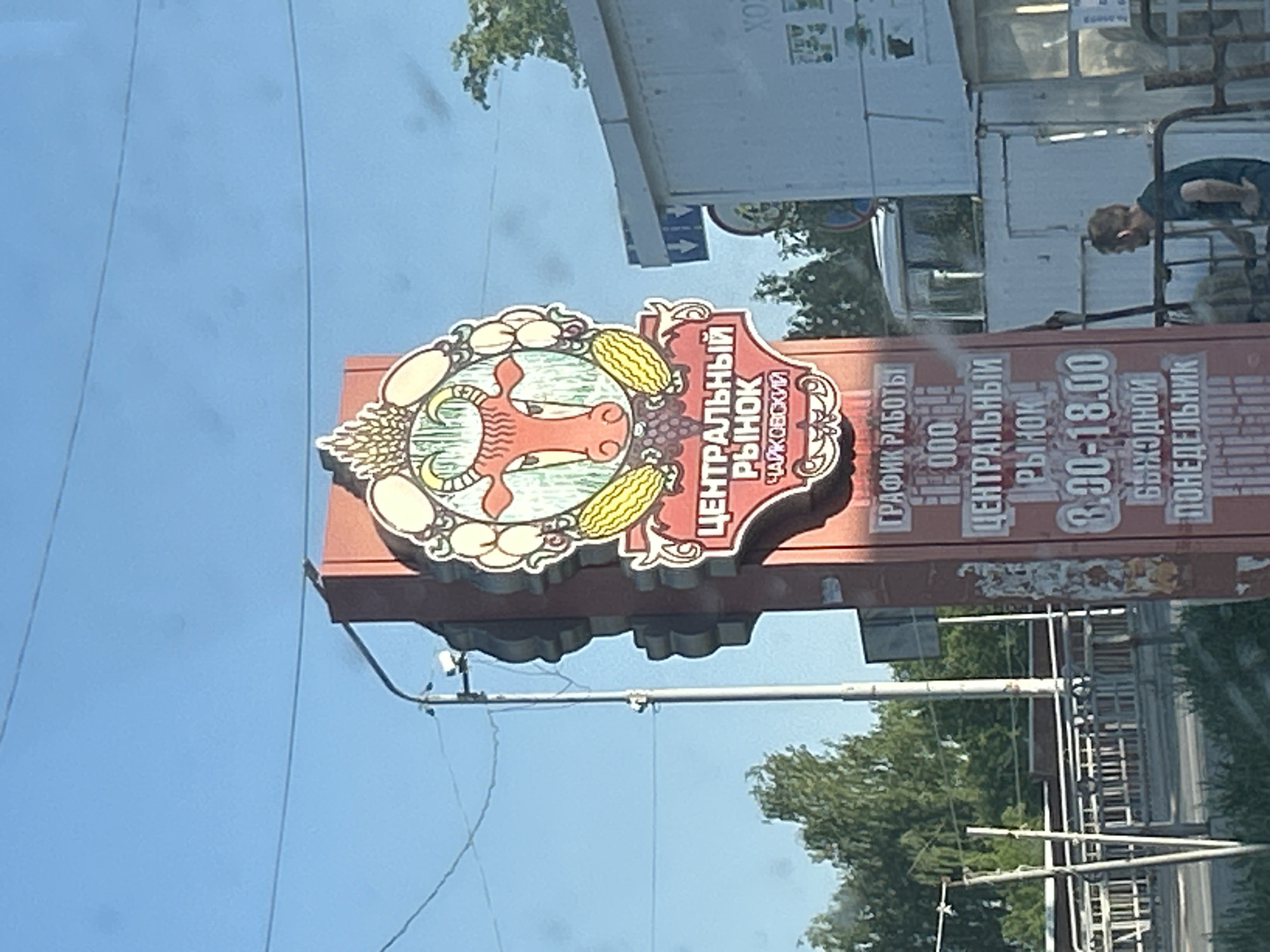 A street-level outdoor view depicts a sign for the "Central Market" in Chaikovsky, Russia. The sign is rectangular and red with white Cyrillic lettering and features a decorative emblem of a cow's head surrounded by images of fruits and vegetables. The text below reads: "Central Market Chaikovsky." The sign also has information about the market's hours of operation: from 9:00 to 18:00 every day, except Monday. A light-colored pole supports the sign. In the background, there's an out-of-focus view of a street, buildings, trees, and a man standing near scaffolding. The sky is visible in the upper left corner and is light blue.