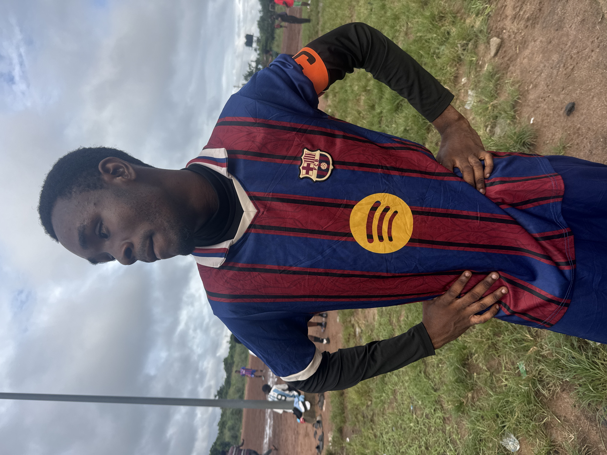 A medium shot captures a young Black man, appearing to be in his late teens or early twenties, standing with his hands on his hips. He is wearing a blue and red striped soccer jersey with a black long-sleeved undershirt. The jersey features the FC Barcelona crest on the left chest and a prominent yellow Spotify logo in the center. An orange armband is visible on his right upper arm. He is looking to his left with a neutral expression.
The background suggests an outdoor setting, likely a soccer field, with dirt and patchy grass. In the distance, other individuals can be seen, some wearing soccer attire. A tall, thin pole stands to the left of the man. The sky above is overcast with clouds. The overall impression is that of a casual sporting event in a somewhat undeveloped area. This scene is located in the city of Ihiagwa, Nigeria.