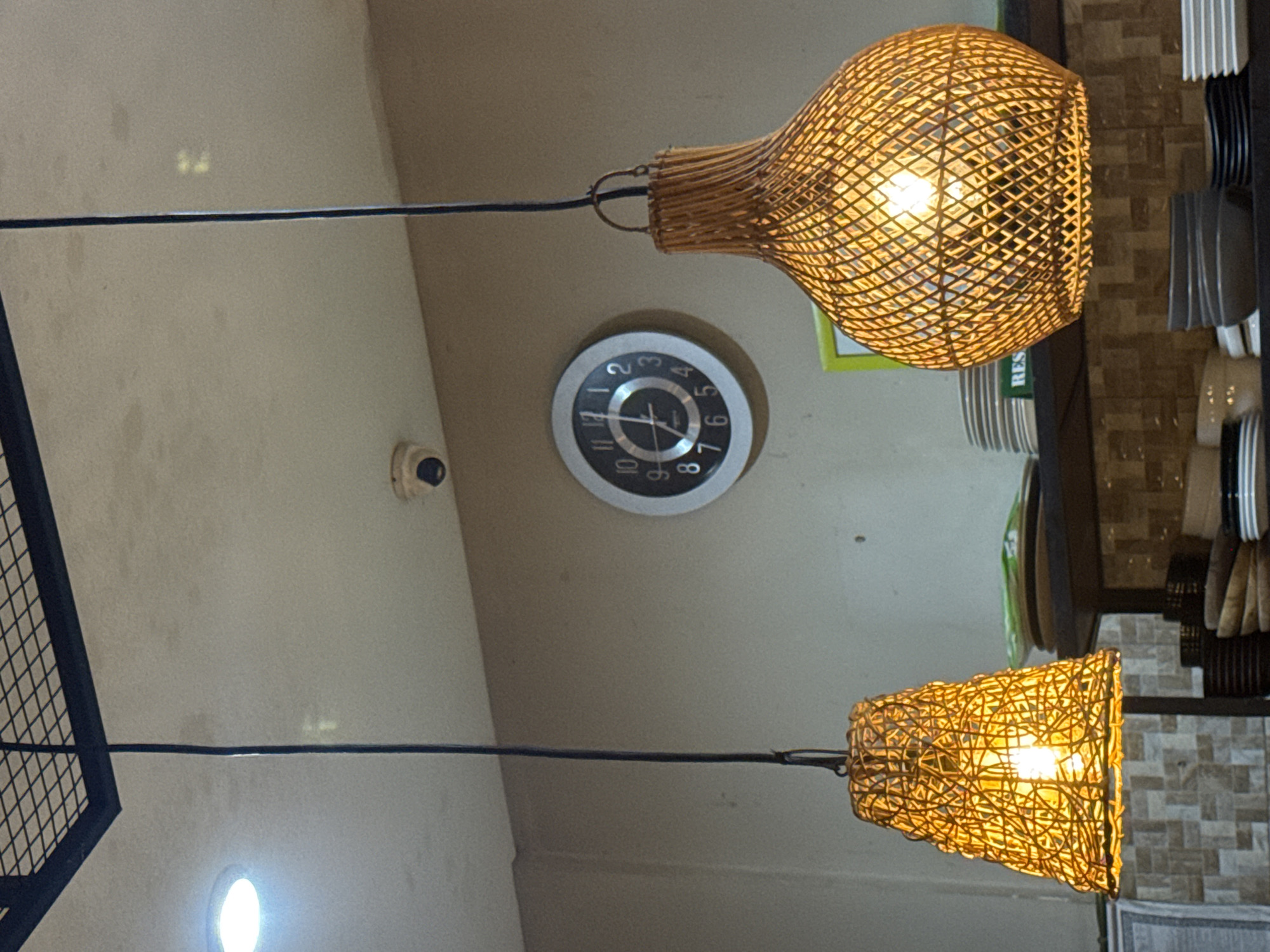The image captures an upward view of an indoor ceiling space, likely within a restaurant or cafe in Kuta Selatan, Indonesia.
**Subjects:** The primary subjects are two illuminated pendant lamps with intricately woven wicker shades, emitting a warm, golden light. A circular wall clock is centered between them. Below the clock, a small, white, dome-shaped security camera is mounted on the wall. To the far left, a section of a black metal grid structure is visible. On the right, shelves display stacks of white and dark-colored plates and cutlery, set against a backdrop of light brown and grey textured wall tiles.
**Activity or event taking place:** The image presents a static indoor scene, focusing on the fixtures and decor of what appears to be a dining establishment. No people or specific activities are occurring.
**Notable details:** The warm glow from the wicker lamps creates a cozy atmosphere. The wall clock indicates the time is approximately 2:10. Faint green text, partially visible as "RES" (likely part of "RESTAURANT" or "RESERVED"), can be seen on a label behind the upper right lamp. The ceiling and visible wall areas are a neutral, light tan color. A bright circular light source is present in the bottom-left corner. The style of the lamps hints at a tropical or natural aesthetic, fitting for the Kuta Selatan location.