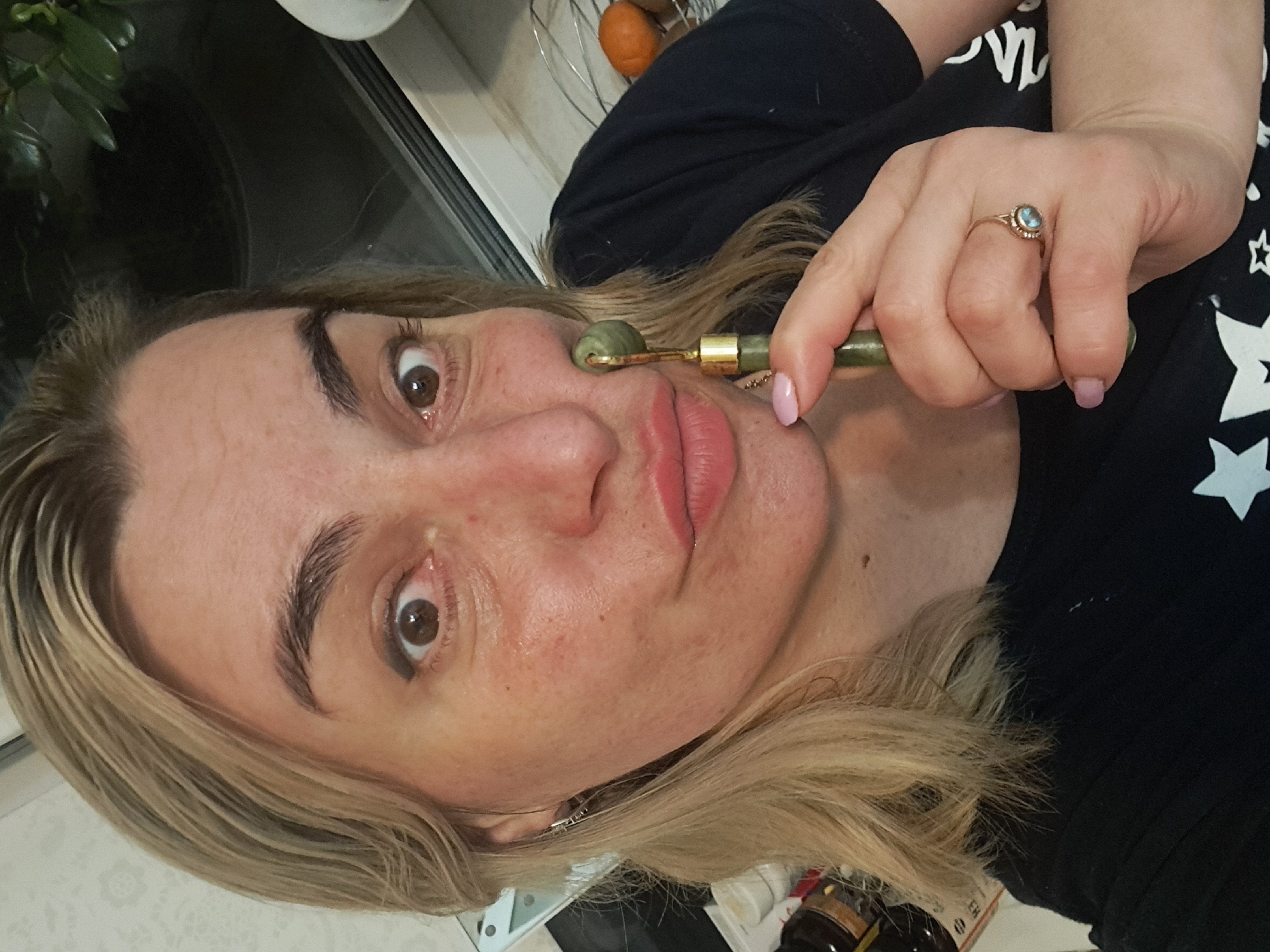 A woman is facing the camera, holding a green jade roller to her face. She has blond hair, fair skin, and brown eyes. She is wearing a ring on her right hand and a black shirt with white stars. The background includes a window, various objects on a countertop, and what appears to be an orange in a wire basket. The location, Elektrostal, Russia, cannot be confirmed from visual data.