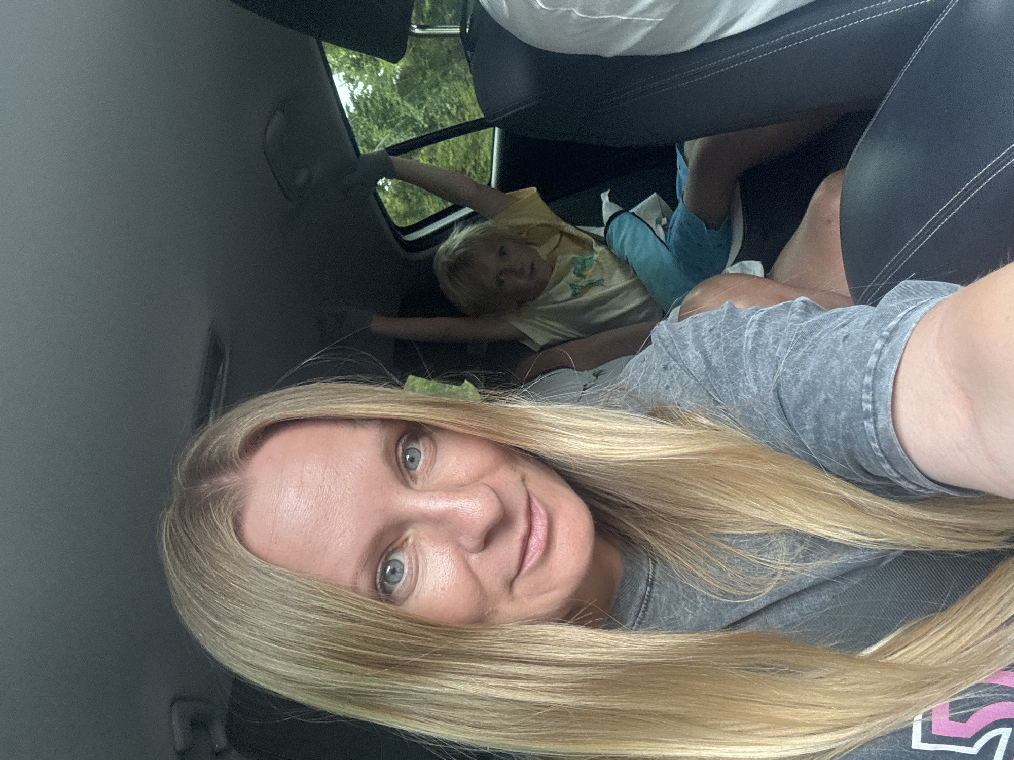 A selfie taken inside a car shows a woman with long blonde hair and blue eyes in the foreground, smiling at the camera. She is wearing a gray t-shirt. In the background, a young child with blonde hair is sitting in the back seat with their arms raised, wearing a yellow t-shirt and blue shorts. The car interior is visible, including car seats and the window showing trees and foliage outside, suggesting a rural or natural setting near Šavnik, Montenegro. The overall scene conveys a casual, everyday moment during a car journey.