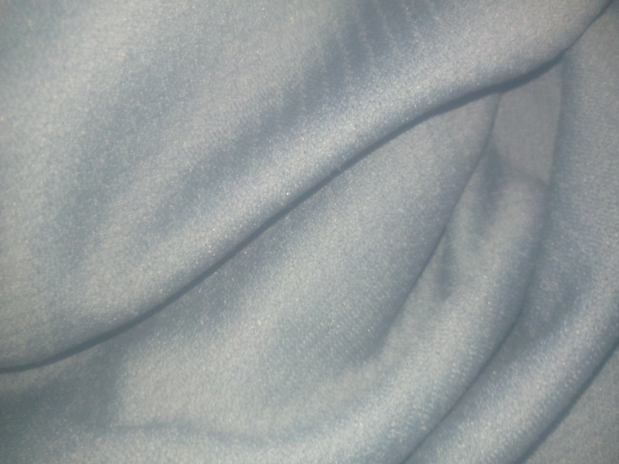 The image is a close-up, abstract shot of a light blue, textured fabric. The fabric is draped and folded, creating soft curves and shadows. There are no people, objects, or environmental details visible that would allow for an identification of its location within Jauro Yinu, Nigeria, or any other city. The focus is solely on the texture and folds of the material.