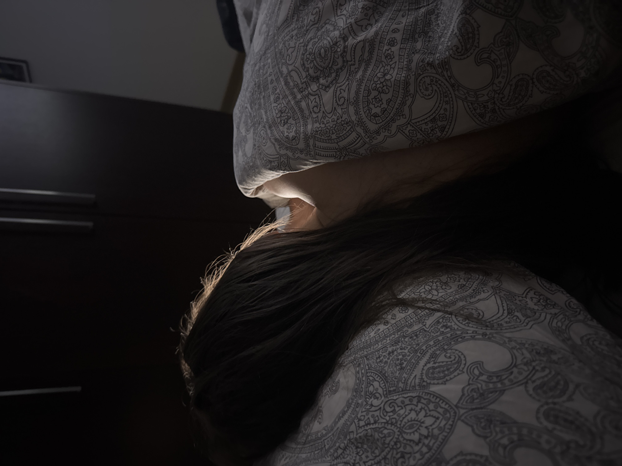 A person is lying in bed with their head resting on a pillow patterned with a white and gray paisley design. The person's dark hair is visible, and their face is obscured by the pillow. A hand rests against the pillow near the person's face. In the background, a dark-colored dresser with silver drawer pulls is partially visible. The lighting is dim, with a light source positioned above the person, casting shadows in the room. The image appears to be taken indoors in a bedroom. The location in the city of Warsaw, Poland cannot be determined from the image alone.