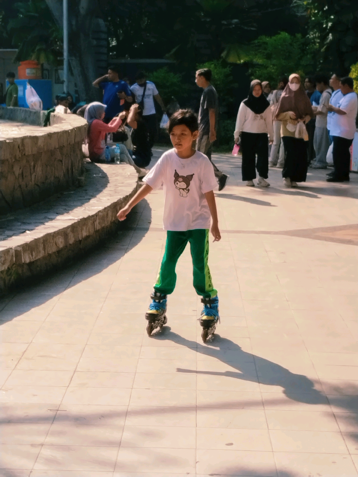 In Surabaya, Indonesia, a young boy with dark hair and a light complexion is rollerblading on a tiled pathway. He is wearing a white t-shirt with a graphic print of a character resembling a dog and green pants with a yellow stripe. His rollerblades are blue and black. He is in motion, with his right arm extended behind him, and his shadow stretches across the tiled ground. In the background, a stone wall curves along the left side of the pathway, with people gathered behind it. Several individuals are visible, including women wearing headscarves and men in casual attire. Trees and foliage create a natural backdrop. The lighting suggests it is daytime, with strong sunlight casting distinct shadows.