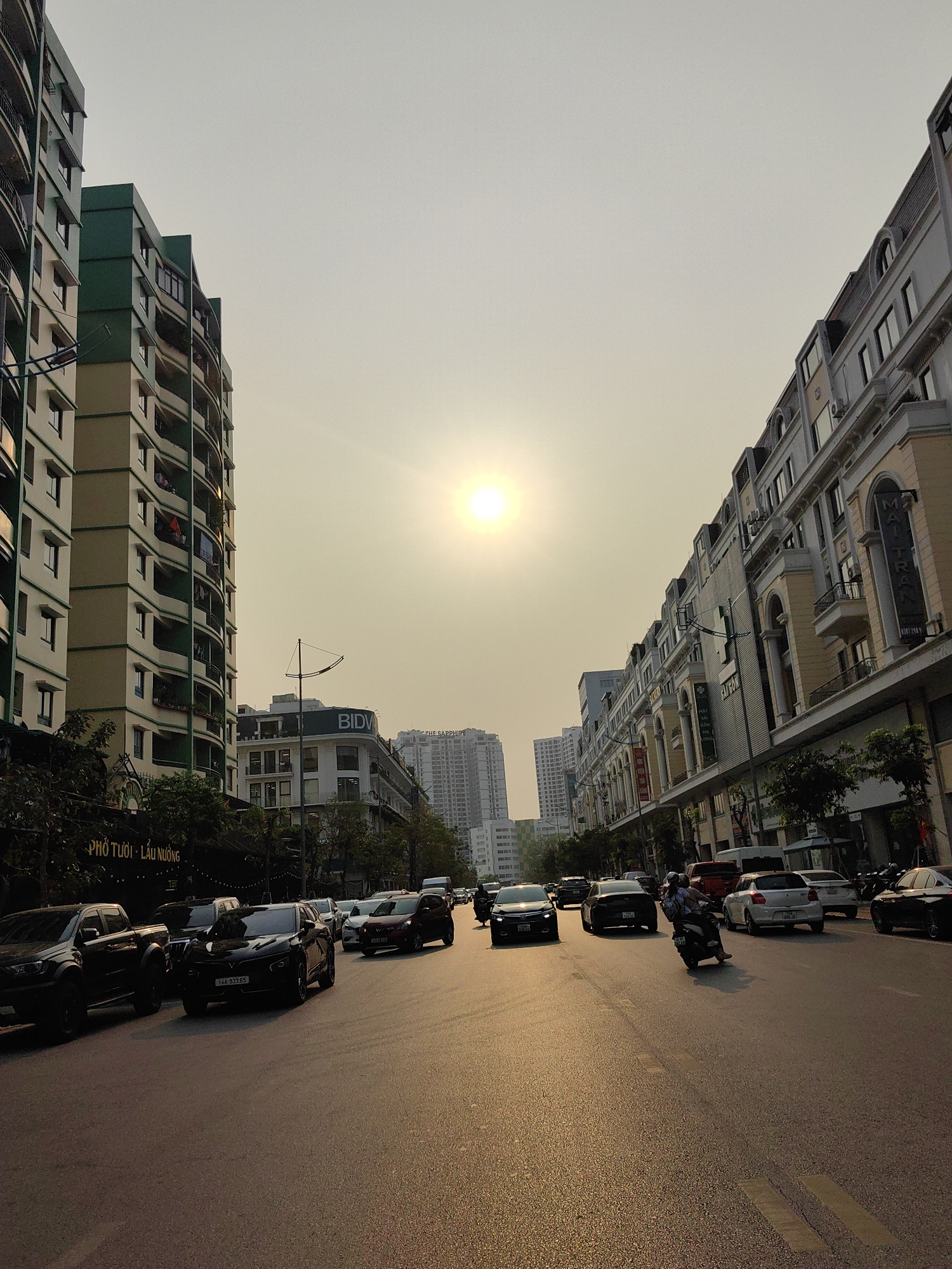Captured from ground level, the photograph presents a daytime urban street scene. It appears to be a wide asphalt thoroughfare bustling with vehicular traffic, including cars and motorcycles. A lone scooter, with two riders, maneuvers along the road's center.
The street is lined with buildings of varying architectural styles and heights. The structure on the left exhibits a modern design, with a light green facade and protruding balconies. On the right, the buildings display a more ornate, classical design, featuring arches and decorative elements. Storefronts are visible on the ground floors of the buildings.
The sky is a uniform, pale yellow, indicating possible atmospheric conditions, like haze. The sun is visible, and appears to be low in the sky, casting long shadows across the road.
Based on the signage visible, the photograph shows elements common to city of Hong Gai Ward, Vietnam.