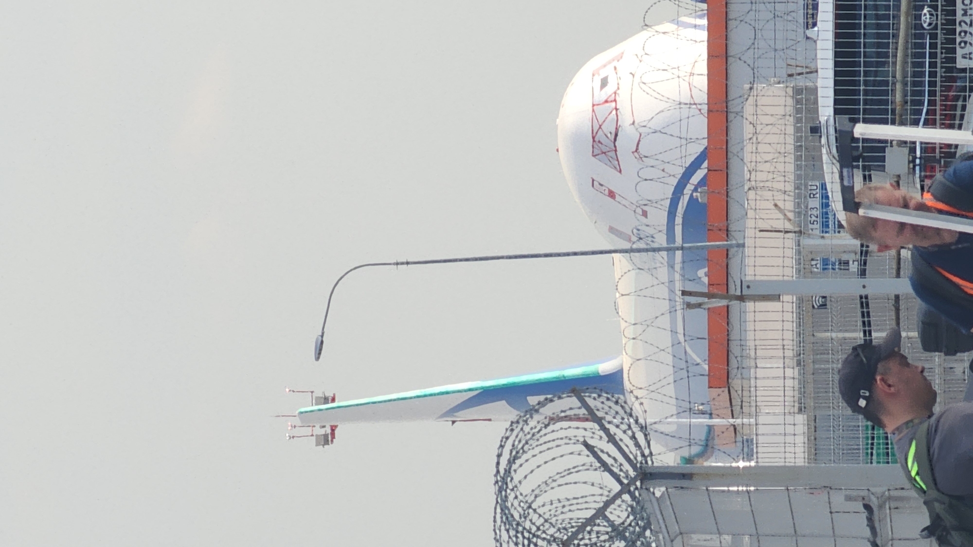 The image shows part of an aircraft, specifically the tail section with a white, blue, and orange color scheme, located behind a barbed wire fence. The tail fin has a green and white stripe. Visible markings on the aircraft include "1523 RU." A light pole is positioned near the aircraft. The environment appears to be an outdoor area with a hazy sky. Two men are partially visible on the right side of the image, standing near the fence. Based on the available information and visual elements, a precise location within Khimki, Russia, cannot be determined with certainty. However, the presence of an aircraft suggests an airport or aviation-related facility.