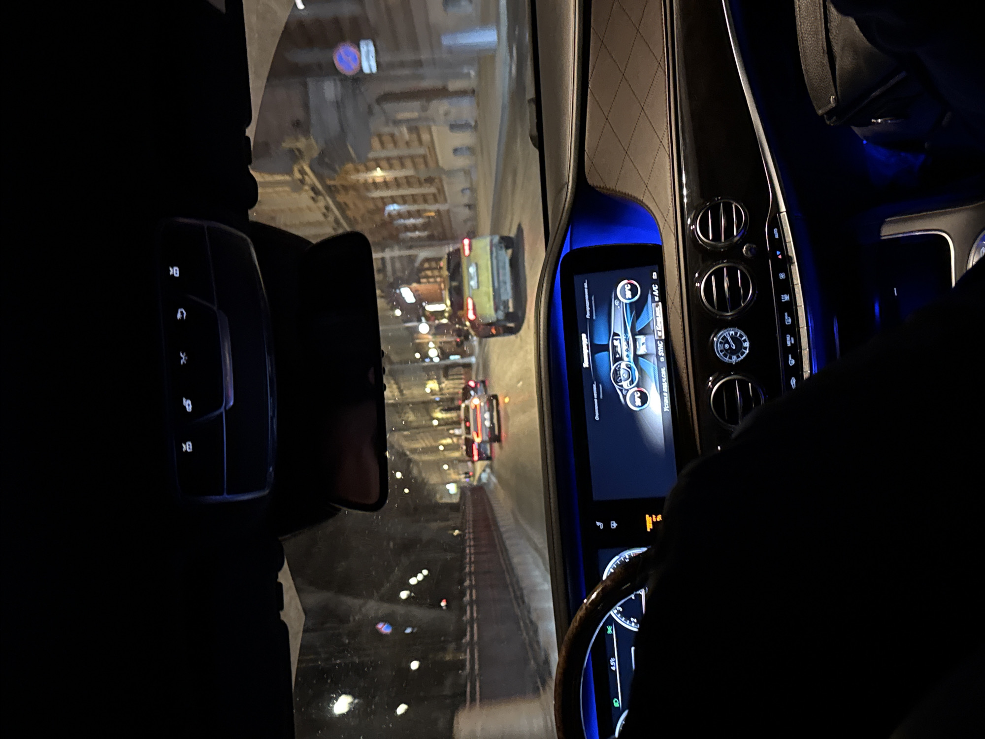 The image, taken from inside a car, shows a view of a street at night in Moscow, Russia. The car is stopped in traffic. Various vehicles, including a yellow bus, are visible through the windshield. The interior of the car features a digital display screen, air vents, and a steering wheel. Several control buttons and indicators are also visible on the dashboard. The ambient lighting inside the car is blue.
