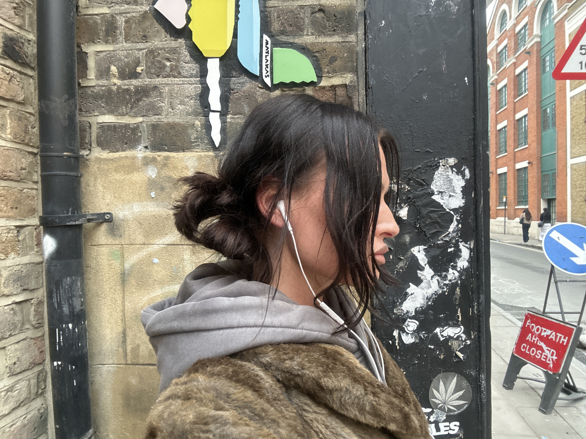 A person is depicted in profile, facing right, standing on a street in what appears to be a metropolitan area of London, United Kingdom. The individual has dark brown hair, styled in a messy bun at the back with strands loose around the face, and is wearing white wired earbuds. They are dressed in a grey hooded sweatshirt under a brown, mottled faux-fur coat.
To the immediate left of the person is a beige and dark brick wall with a vertical black drainpipe secured to it. Above the person's head, on the brick wall, are colorful abstract mosaic art pieces, including yellow, light blue, and green segments, with "MAATALAS" vertically inscribed on one blue piece.
To the immediate right of the person is a tall, black, textured pillar or post, displaying evidence of peeling white paper and stickers, including a white cannabis leaf sticker near the bottom.
In the background, to the right, a city street is visible with light grey pavement and asphalt. Two pedestrians are discernible further down the street. A prominent red and white triangular road sign with a black "5" over "10" is visible, indicating speed limits or other road information. Below it, a blue circular sign with a white arrow pointing diagonally downwards to the right suggests traffic flow or a bypass. Closer to the foreground on the street is a red A-frame sign with black text reading "FOOTPATH AHEAD CLOSED," indicating a pedestrian path closure. Beyond the street, a large multi-story building constructed of red brick with numerous tall, arched windows and green-framed window sections on its facade is present. The architecture is consistent with older industrial or warehouse buildings common in London.