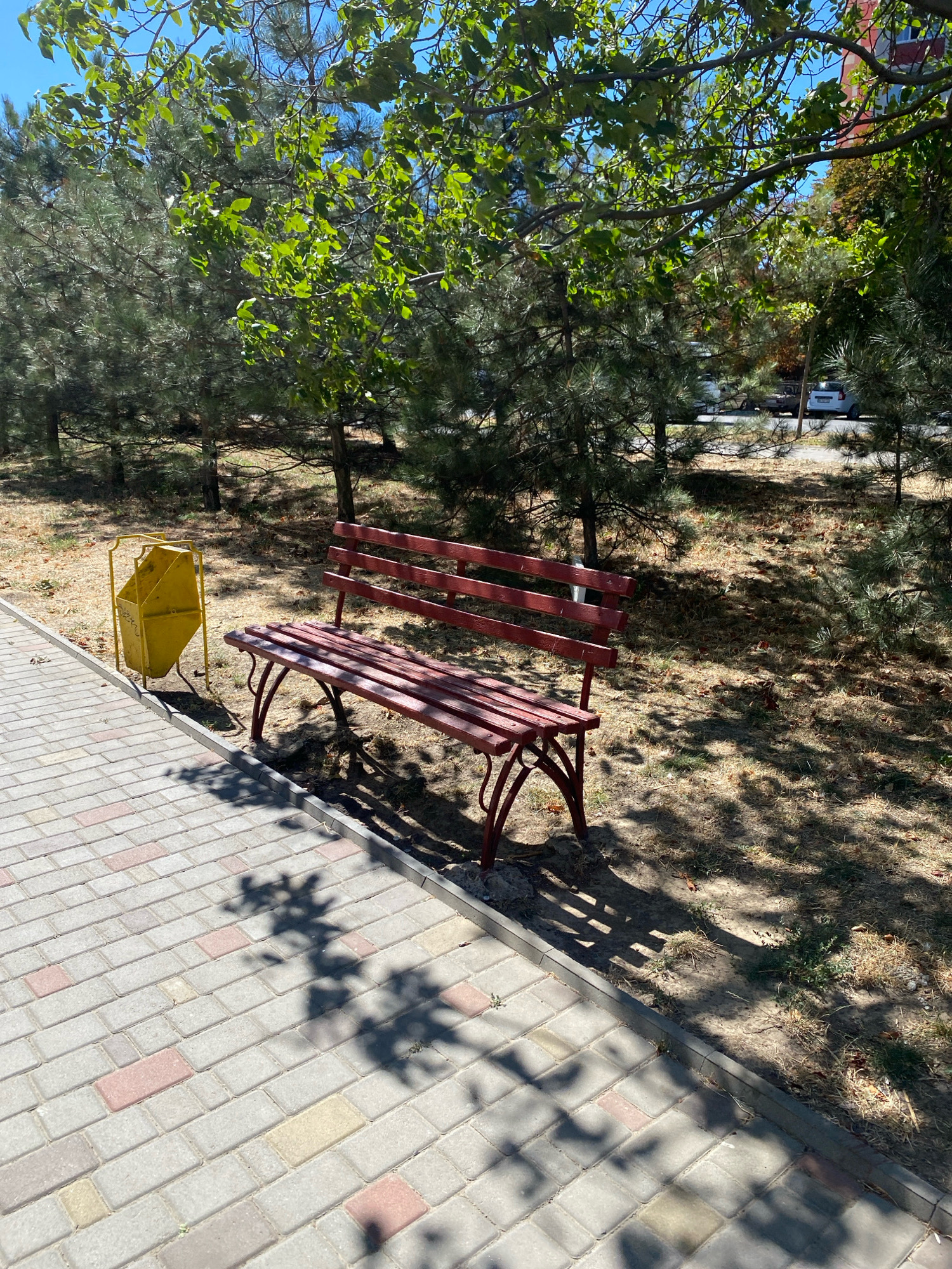 The image depicts a park scene in Chornomorsk, Ukraine, under a clear blue sky. In the foreground, a paved pathway made of light-colored interlocking bricks, some with pink and yellow accents, runs diagonally from the bottom left to the top right. To the right of the pathway is a strip of dry grass and dirt. A red metal park bench with wooden slats is positioned on the dirt area, facing left, with its shadow cast on the ground. To the left of the bench and slightly further back is a yellow metal trash can with an angled opening. Behind the bench, a line of mature pine trees with dense needles creates a natural screen. Further back, more trees with green foliage are visible, and in the distance, cars can be seen on a road, along with the corner of a red building. The lighting suggests it is a bright, sunny day, with dappled shadows from tree branches falling across the pathway and ground.