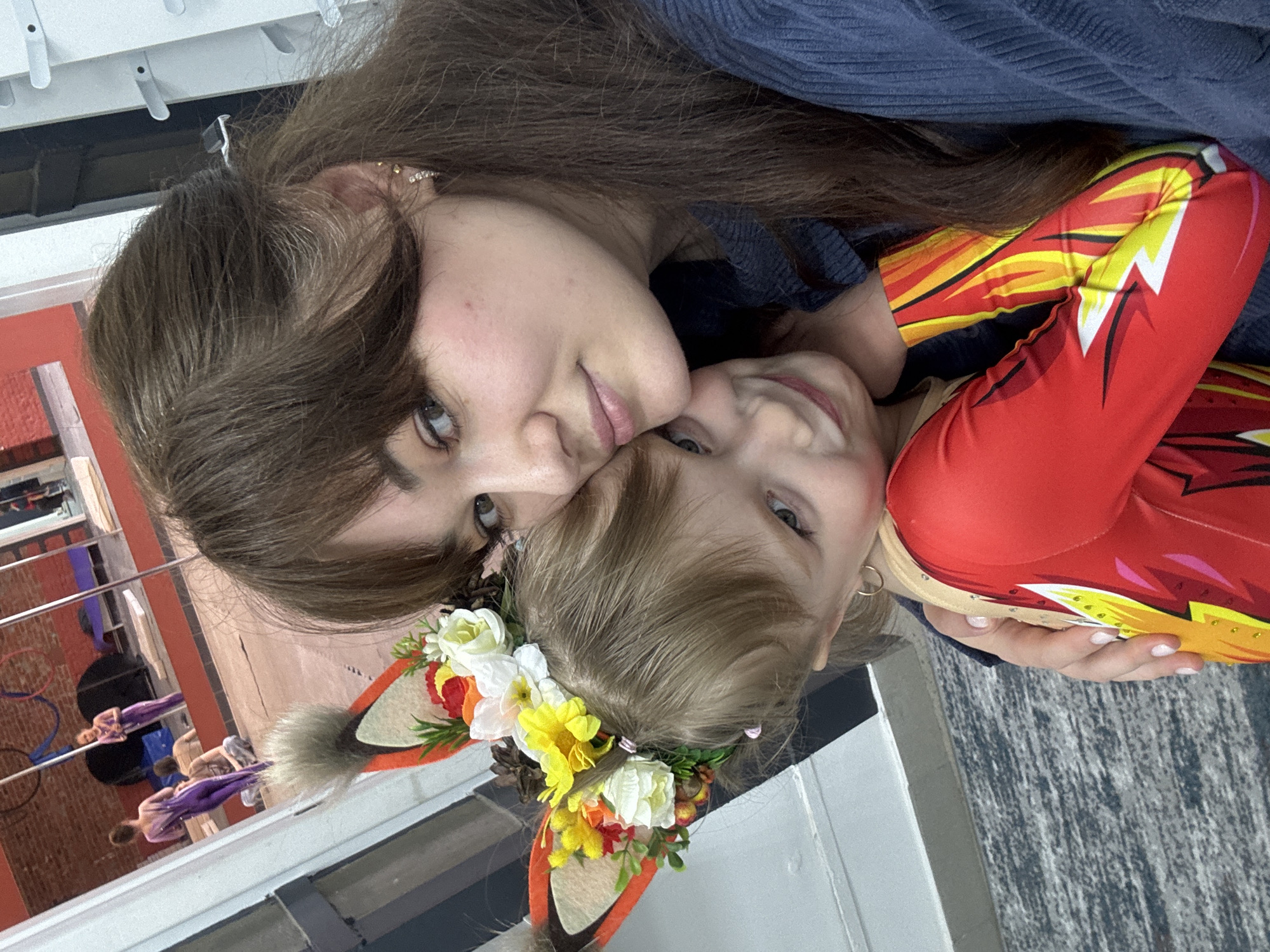 A close-up, vertically oriented photograph captures an adult and a child in an affectionate embrace within an indoor setting in Chaikovsky, Russia. The adult, positioned towards the top of the frame, has long brown hair and dark eyes, looking directly at the camera with a gentle, contented expression. She wears a dark blue ribbed top and her left ear is adorned with multiple small, shiny earrings.
The child, with lighter hair and eyes, is nestled closely against the adult, looking slightly upwards with a cheerful smile. They are dressed in a vibrant red and yellow outfit, possibly a leotard or jumpsuit, featuring a prominent white lightning bolt design, suggestive of a superhero costume. The child wears an elaborate headband with faux animal ears, decorated with a wreath of artificial flowers in white, yellow, orange, and red, interspersed with small berries or pinecones. A simple gold hoop earring is visible on the child's left ear.
The background, partially blurred, reveals an indoor facility, likely a gymnasium, studio, or performance space. To the left, metal rigging and a red wall are discernible. In the deeper background, several blurred figures in purple outfits appear to be engaged in aerial arts or gymnastics, indicating an active environment. The floor has a textured, possibly speckled, surface. The overall mood of the image conveys warmth and happiness, highlighting the close bond between the two individuals. There is no visible text in the image.