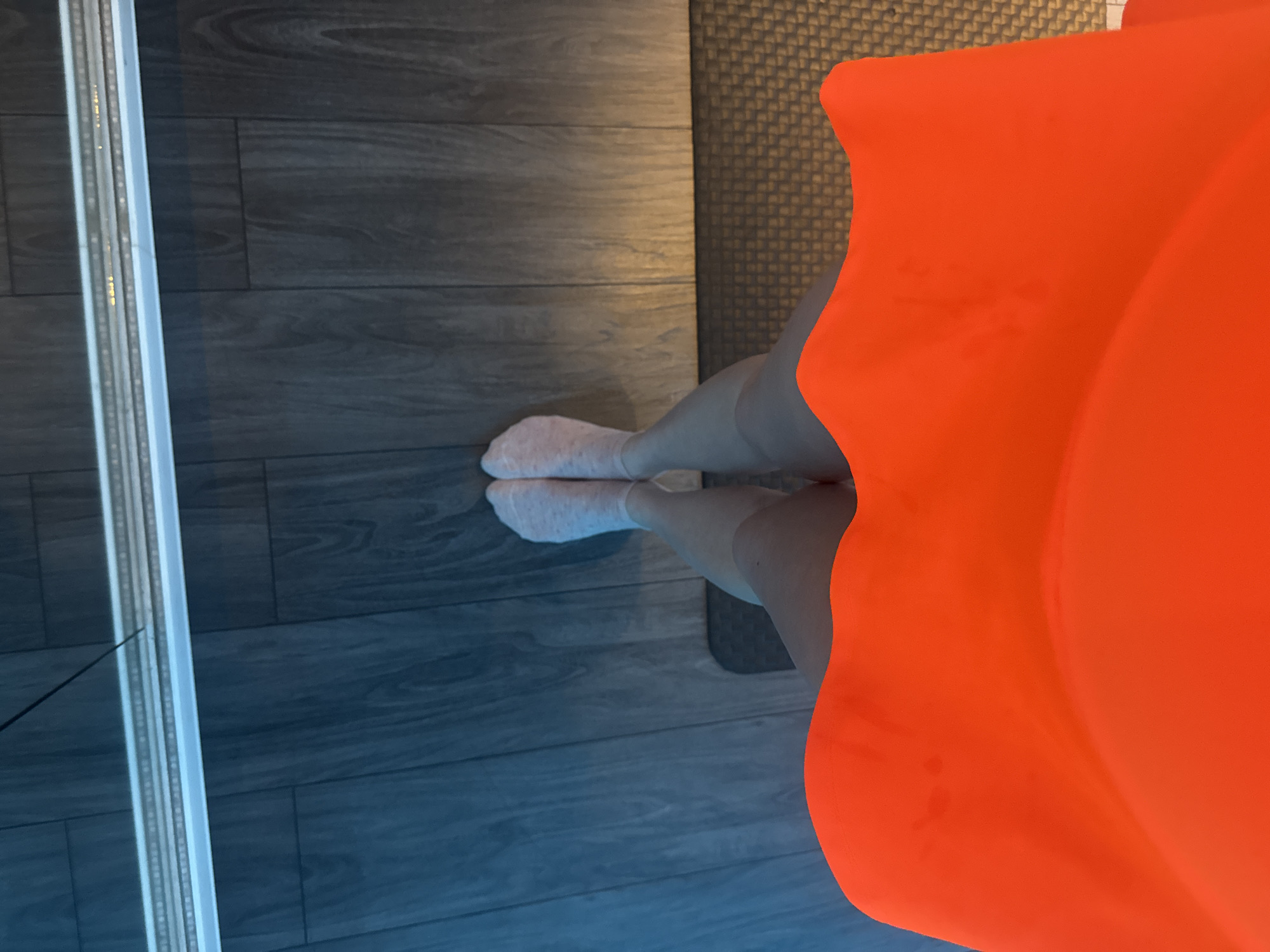 A top-down shot shows a person standing indoors. They are wearing white socks and an orange garment that is cut off at the waist. The person is standing on a surface that transitions from gray wood flooring to a dark gray textured mat. A metal strip runs along the left edge of the frame. The location is Unknown, Unknown.