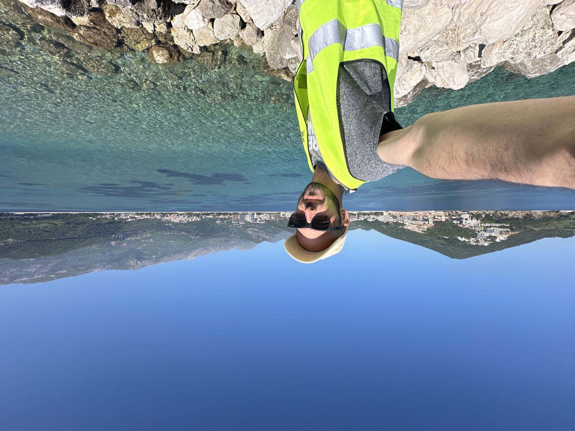 A man in a yellow safety vest, sunglasses, and a hat is positioned in the foreground. The background consists of rocks and the sea. In the distance, there is a view of the city of Budva, Montenegro, with mountains visible behind the city. The image is rotated 180 degrees.