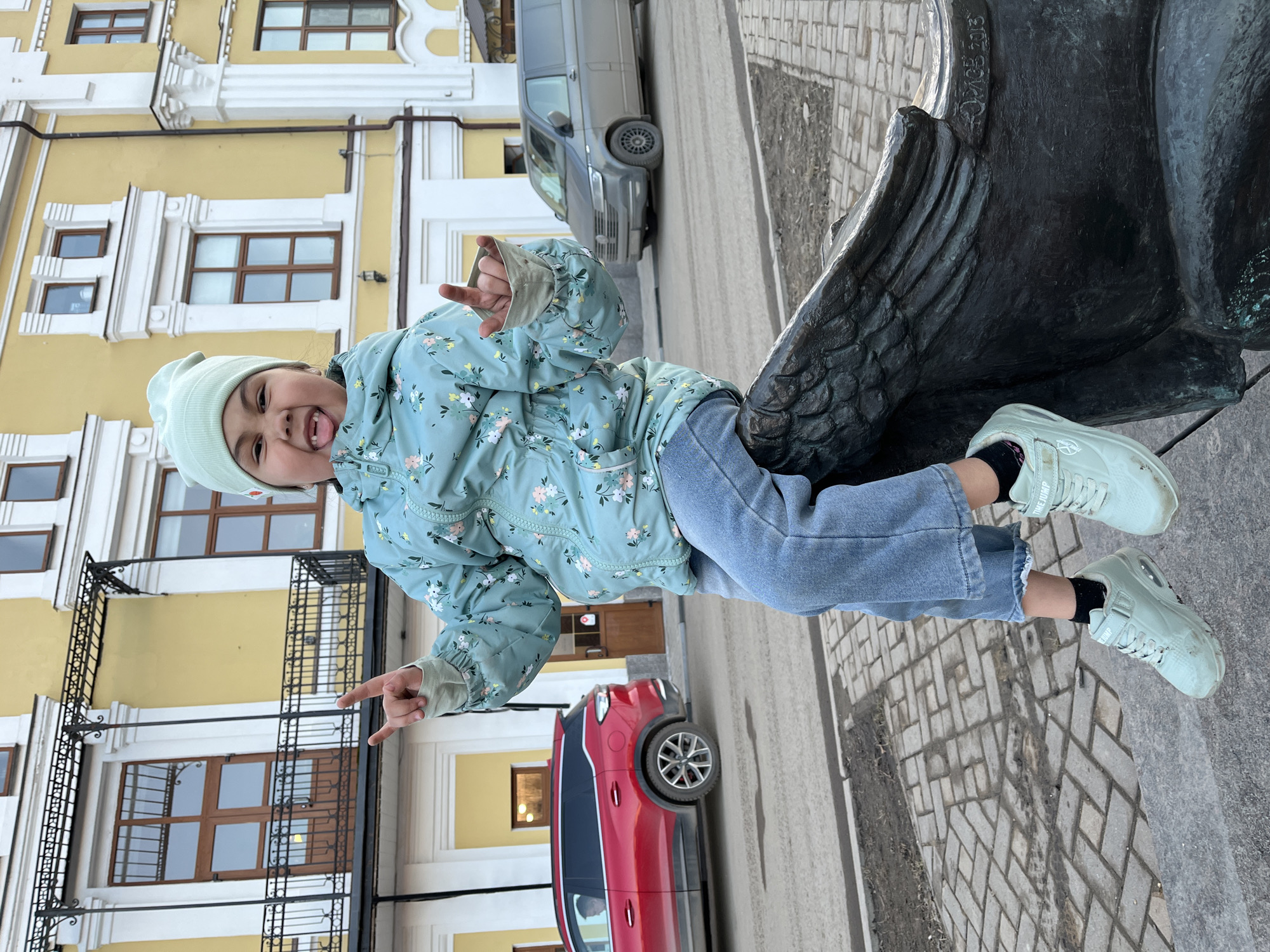 A young child is positioned on a statue in Podolsk, Russia, as evidenced by "OMCB 2013" etched on the statue. The child is wearing a light blue patterned jacket, jeans, a light green beanie, and light green sneakers with black socks. The child is sticking their tongue out and making hand gestures. In the background are a yellow building with brown-framed windows, a red car, a grey car, and a cobblestone walkway.
