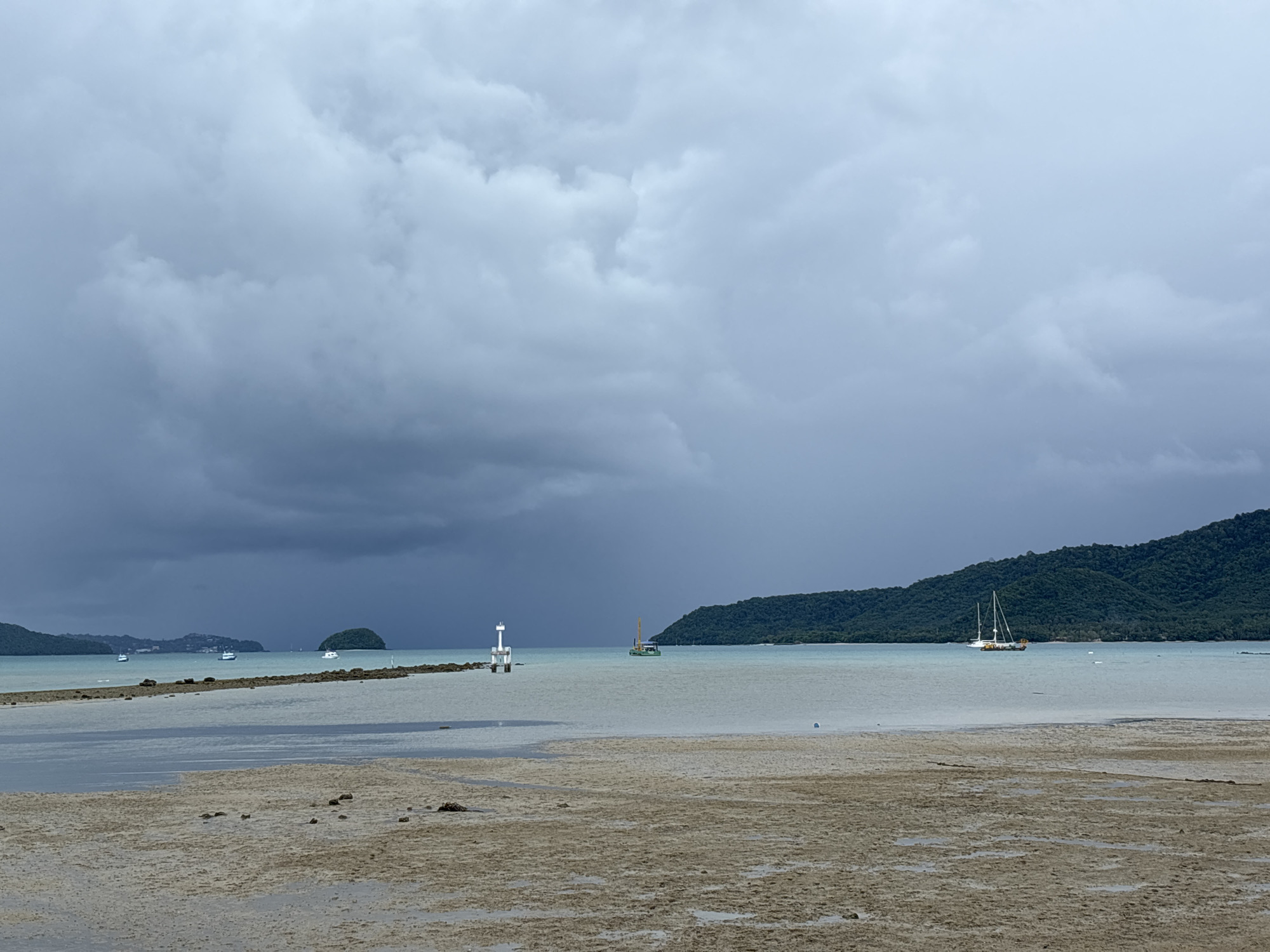 A serene, overcast scene captures a tranquil bay in Rawai, Thailand. In the foreground, a sandy, wet shore is dotted with scattered rocks and shells. The middle ground features a shallow, calm body of water with a visible rocky outcrop extending from the left. A white and red navigation marker stands on this outcrop, guiding vessels. Further out, several boats are moored in the bay. Two white sailboats are prominent on the right, one larger with two masts and a yellow accent, the other smaller. Another small white boat is visible in the distance near the center.
The background is dominated by a lush, green, tree-covered hill that curves around the bay. The sky is heavy with dark, dramatic storm clouds, particularly on the left side where a faint rain shower is visible. The overall atmosphere is subdued and peaceful, with the overcast sky creating a soft, diffused light.