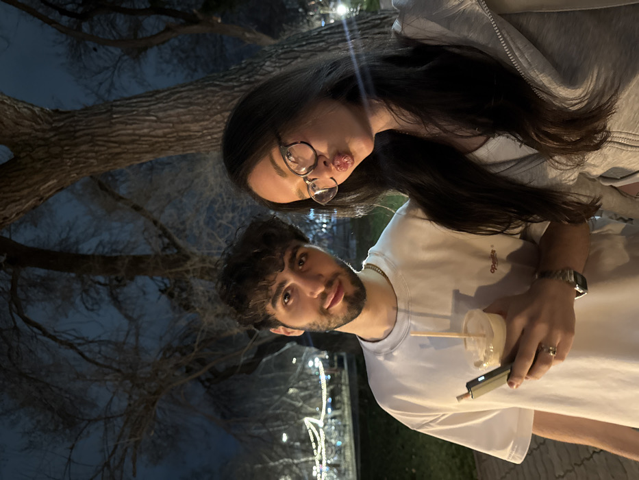 Two individuals, one male and one female, are depicted in a close-up, waist-up portrait taken at night in a public park or pedestrian area in Yerevan, Armenia.
The male subject, positioned on the left, has dark, curly hair and a short beard. He wears a light-colored crew-neck t-shirt with a small dark "operātis" logo on the right chest, a gold-colored chain necklace, a ring on his left ring finger, and a watch on his left wrist. He holds a clear plastic cup with a light-colored beverage and a wooden straw in his right hand, and a dark-colored electronic device (possibly a smartphone or vape) in his left hand. His gaze is directed towards the camera with a slight smile.
The female subject, positioned on the right, has long, dark hair and wears glasses with tortoise-shell frames. She is dressed in a light-colored (likely grey) zip-up hoodie over a white top. Her lips are puckered in a "kissing" expression, and she is looking slightly to her right, past the camera.
The background is dark, featuring silhouettes of bare tree branches against a dark blue sky. Prominent tree trunks are visible immediately behind the subjects. In the mid-ground and background, blurred string lights and other artificial illumination are visible, suggesting decorative lighting within an urban park setting. A dimly lit paved surface and some grass are visible at the bottom of the frame.