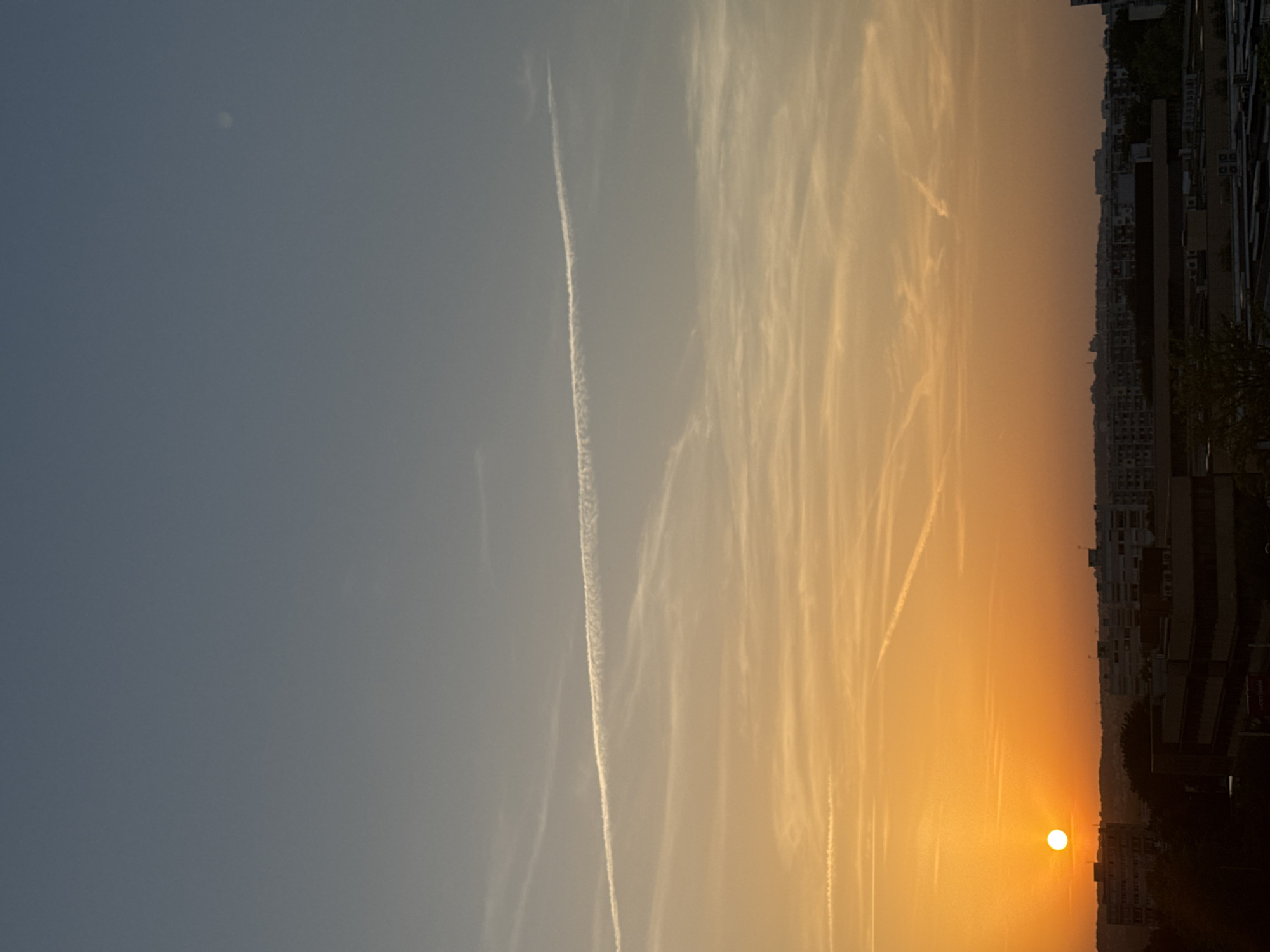 The image shows a city skyline at sunset or sunrise. The sky is a gradient of blue and orange, with several contrails from airplanes streaking across it. The sun, a bright white orb, is visible on the left side of the frame, partially obscured by the horizon. Buildings of various heights and styles are visible in the foreground and mid-ground, with some trees interspersed among them. The overall scene depicts an urban environment under a dramatic sky.