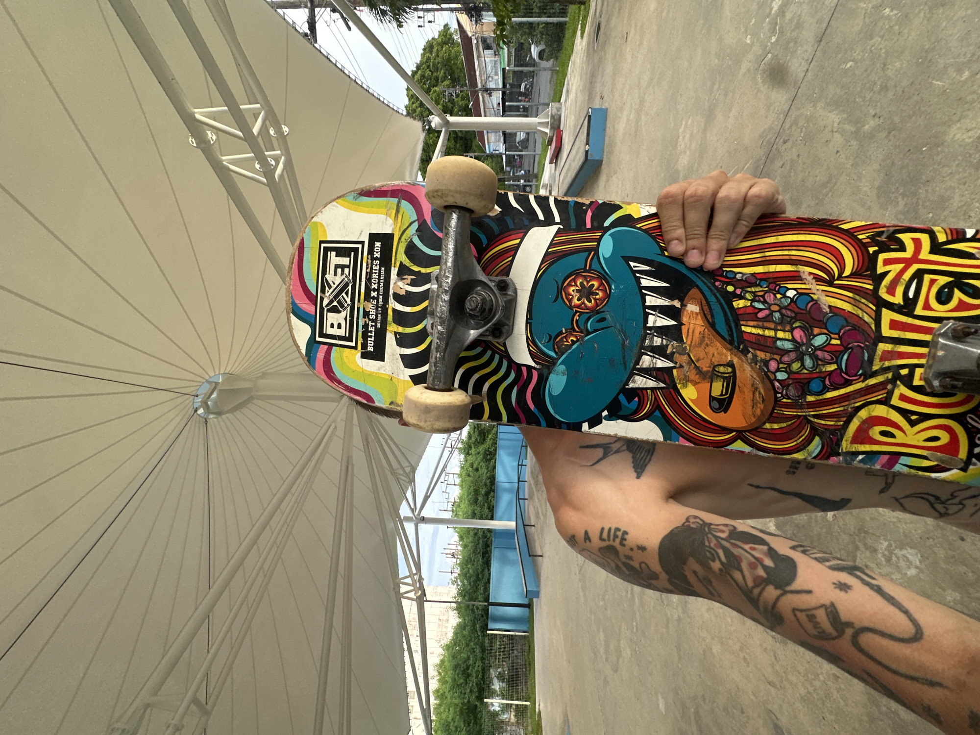 A person, partially obscured by a skateboard, stands in a skate park. The skateboard, held vertically in front of the person's face, displays a colorful, cartoonish graphic with the words "BUXET" and "BULLET SHOE X XORIES XON" printed on it. The person's left arm, visible on the left side of the frame, is covered in tattoos. The skate park features concrete ground, blue ramps, and a white, tent-like shade structure overhead. In the background, trees and buildings are visible, suggesting an urban or suburban setting. The scene is outdoors under what appears to be daylight. The location is within the Phuket City Municipality, Thailand.