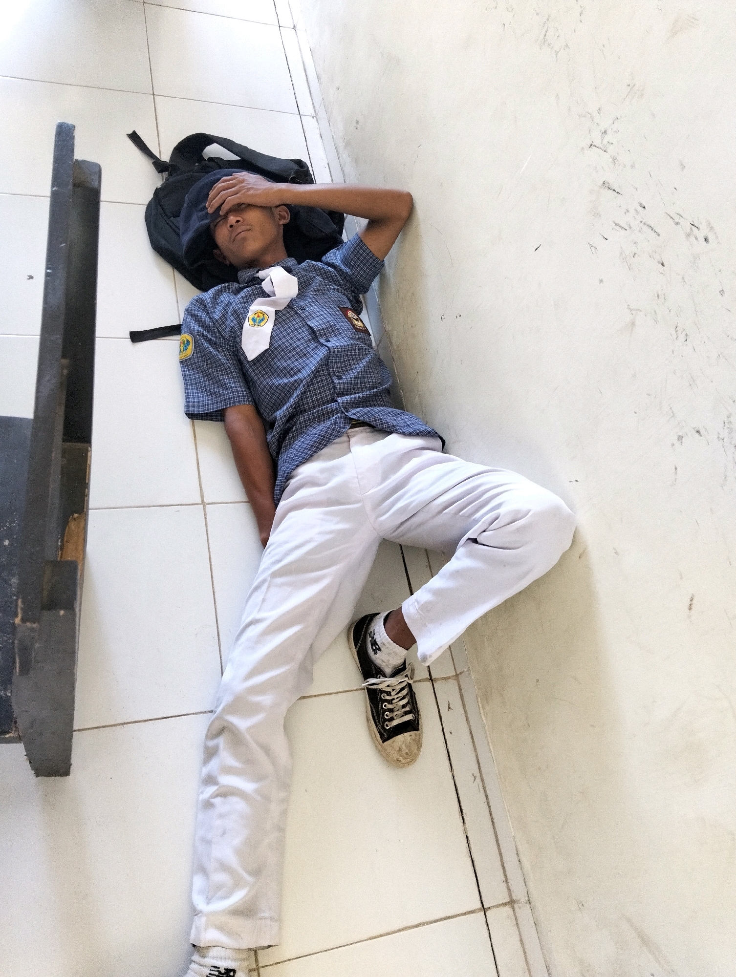 A young male student wearing a blue checkered school uniform shirt and white pants is lying on his back on a tiled floor, with his head resting on a black backpack. His right hand is placed on his forehead, and his eyes are closed. He is positioned near a dark-colored table or bench, with his body extending into a wider tiled area. The background consists of a white wall with some scuff marks. The scene appears to be indoors, possibly in a school hallway or common area in Genteng, Indonesia.