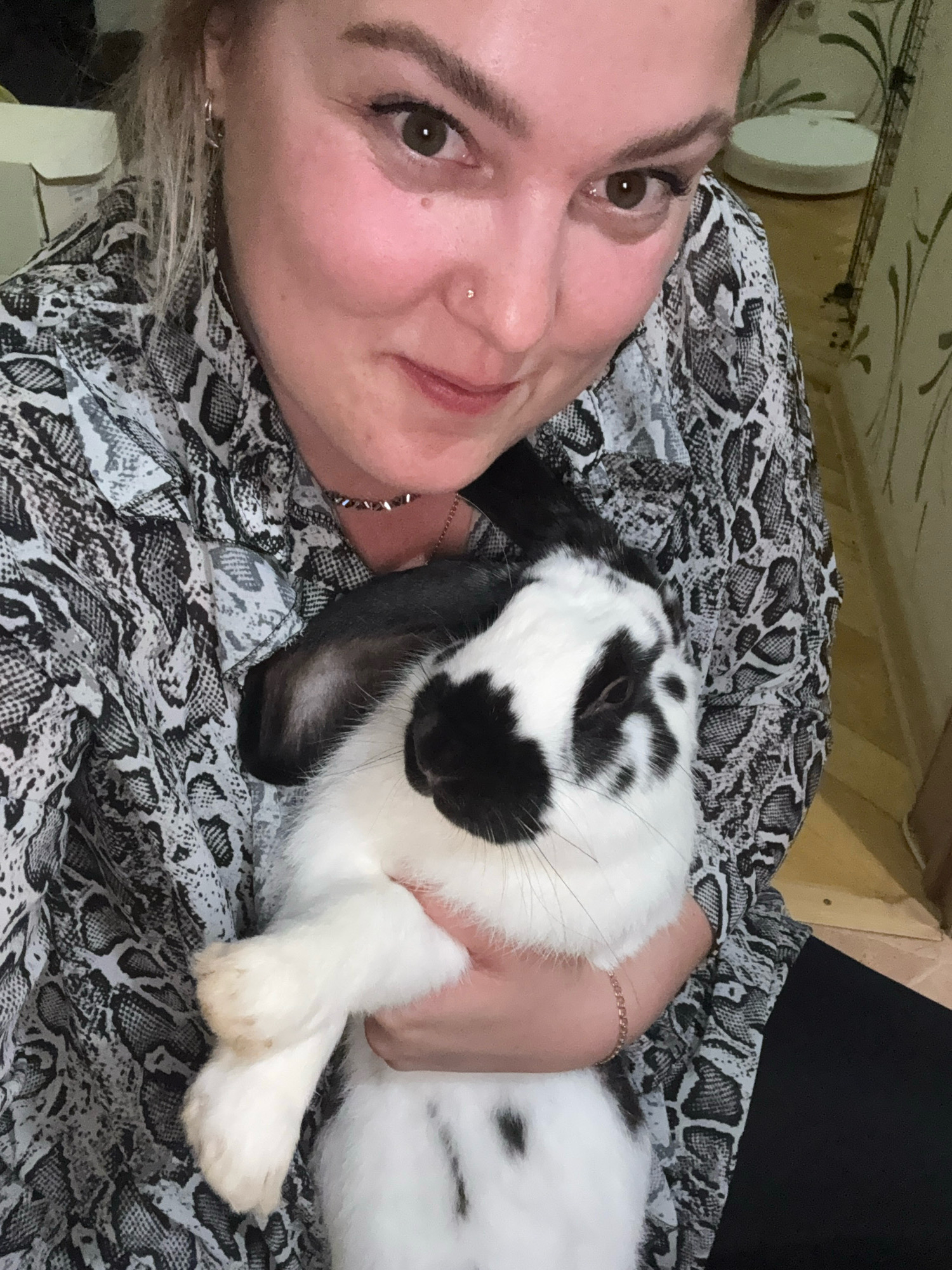 In the city of Марковский, Russia, a young woman with blonde hair and light skin is holding a black and white rabbit. She is wearing a black and white snakeskin-patterned shirt and a thin necklace. The rabbit is nestled in her arms, with its head resting on her shoulder. The woman is looking at the camera with a slight smile, and the rabbit is also looking towards the camera. The background of the image is blurred, but it appears to be an indoor setting with wooden floors and patterned wallpaper. There are no other people or objects clearly visible in the background. The lighting in the image is soft and natural, suggesting it may have been taken during the day. The overall impression is one of a casual and affectionate moment between a woman and her pet.