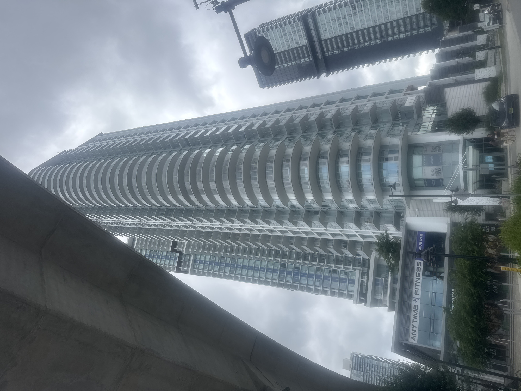 The image depicts a street-level view of a modern urban environment in Coquitlam, Canada, under an overcast sky. Dominating the scene are two tall, contemporary residential high-rise buildings. The building on the left features a distinctive curved facade with numerous balconies, while the building on the right is a more rectangular structure with a dark facade. In the foreground, a low-rise commercial building is visible, with a prominent sign for "ANYTIME FITNESS" on its upper level. Trees and greenery line the street, and a street lamp with a dark fixture is positioned in the middle ground. Two vehicles, a black car and a silver SUV, are parked on the street. Two individuals, one adult and one child, are walking a dog on the sidewalk. The overall impression is of a developing urban area with a mix of residential and commercial spaces.