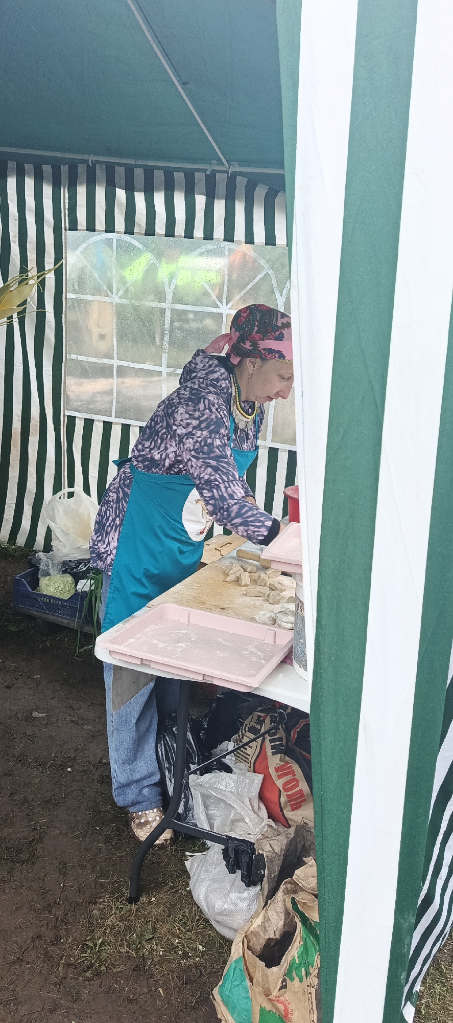 A woman stands behind a table at what appears to be an outdoor market or fair, set up under a green and white striped tent. The woman is wearing a patterned long-sleeved shirt, a bright teal apron, blue jeans, and a pink headscarf with a floral pattern. She is also adorned with a beaded necklace featuring metallic coins. She is actively engaged in preparing food, specifically working with small, dough-like pieces laid out on a wooden cutting board. A pink plastic tray with white powder, likely flour, sits in front of her on the table, and more of the dough pieces are visible on the board.
The setting is outdoors, with the ground appearing muddy and covered in patches of grass. To the left of the woman, a blue crate holds some leafy green vegetables, possibly cabbage, and a white plastic bag. Underneath the table, several bags and what look like trash are piled up. One bag is red with white Cyrillic text that appears to read "УГОЛЬ" (UGOL), meaning "coal." Another bag is brown and green with more Cyrillic text, partially visible.
The lighting suggests it could be daytime, possibly overcast, as there are no strong shadows. The overall mood seems to be one of work and preparation, with the woman focused on her task. The striped tent provides shelter, and the setting suggests a rustic or informal event. The location context of Старые Быги, Russia, implies a local community gathering.