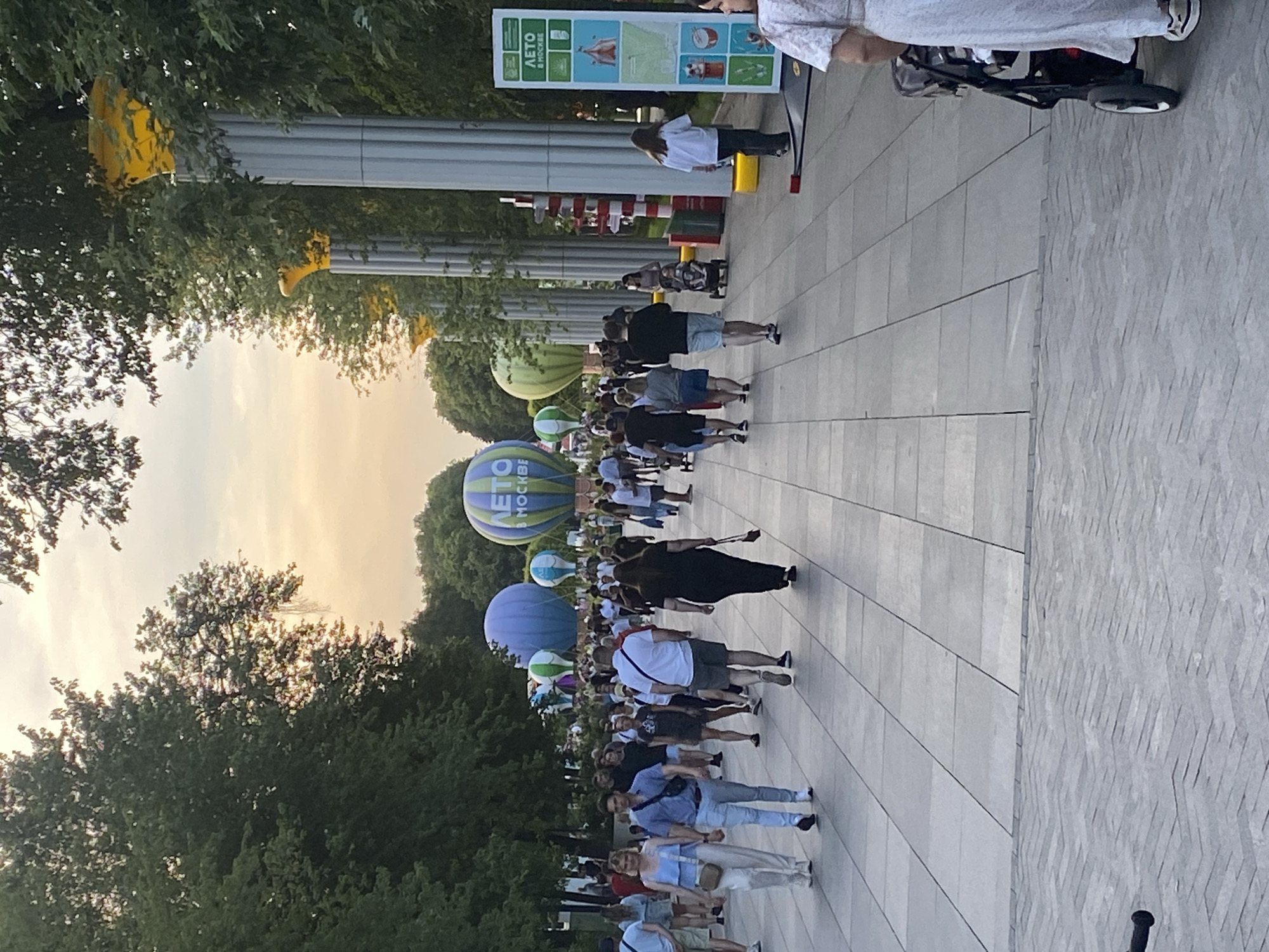 The image depicts a bustling park scene in Moscow, Russia, with a clear sky and a soft sunset glow. The foreground shows a wide, paved walkway with large, square tiles, where numerous people are strolling. To the left, lush green trees line the path, and to the right, large, fluted columns with decorative yellow tops rise above the trees, suggesting an architectural element of the park.
Several hot air balloons are visible in the middle ground and background, some of which appear to be inflated and tethered, while others are smaller and more distant. One prominent balloon features the text "ЛЕТО В МОСКВЕ" (Summer in Moscow), indicating the location and possibly an event. People of various ages are walking, some with children in strollers. They are dressed in casual summer attire. The overall atmosphere is one of a public gathering and leisure activity in an urban green space. A large sign with similar text and graphics is also present on the right side of the walkway.