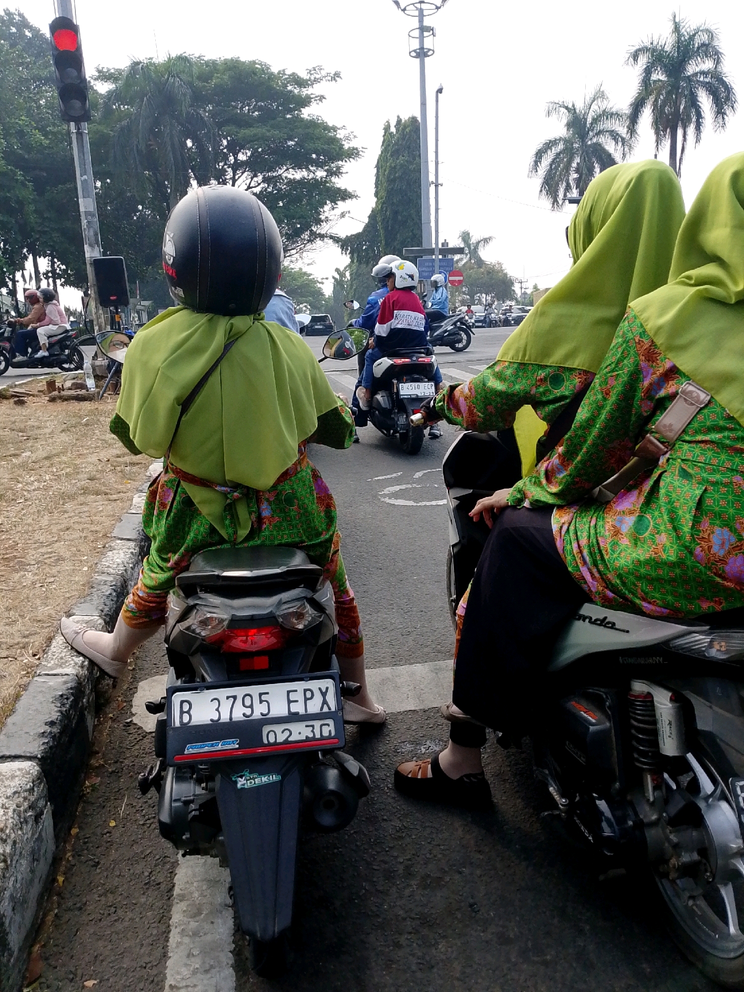 Two women are riding on a motorcycle in Cibinong, Indonesia. The woman in the foreground, wearing a black helmet and a green hijab and dress, is seated on the back of the motorcycle. She is wearing patterned leggings and light-colored shoes. Her right leg is extended. The woman in the background, also wearing a green hijab and dress, is seated in front. The motorcycle has a license plate that reads "B 3795 EPX". Other motorcycles and cars are visible in the background, along with trees and a traffic light. The scene appears to be a street intersection.