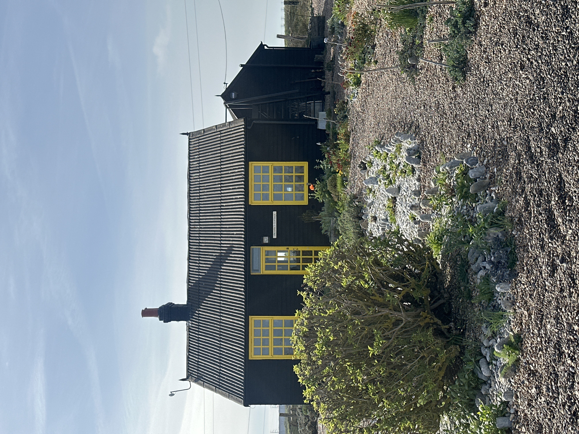 The image depicts a black, one-and-a-half story building with bright yellow window frames and a chimney. The building has a corrugated roof. The surrounding area consists of a gravel path and various plants, including a large bush in the foreground. Electrical lines are visible in the sky. The location is possibly near the coast in the Folkestone and Hythe District, United Kingdom. The building appears to be residential, and the setting suggests a rural or coastal area. The photograph was taken during the day with a clear sky.