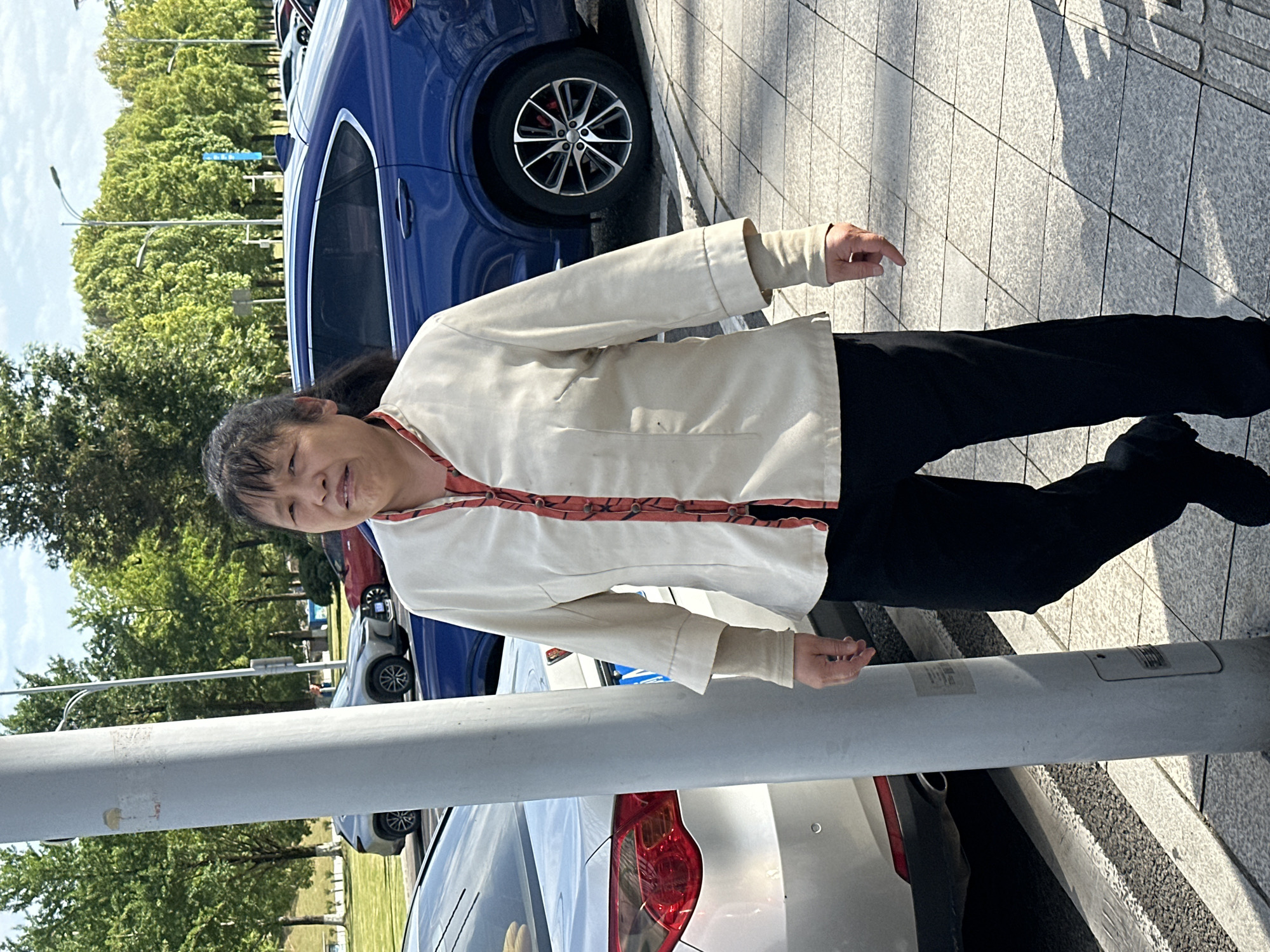 A middle-aged Asian woman stands beside a row of parked cars and a light post. She wears black pants and a light-colored jacket with a decorative trim. She appears to be smiling slightly. The location appears to be Liangjiang, China due to the types of plants and trees as well as style of cars in the shot.