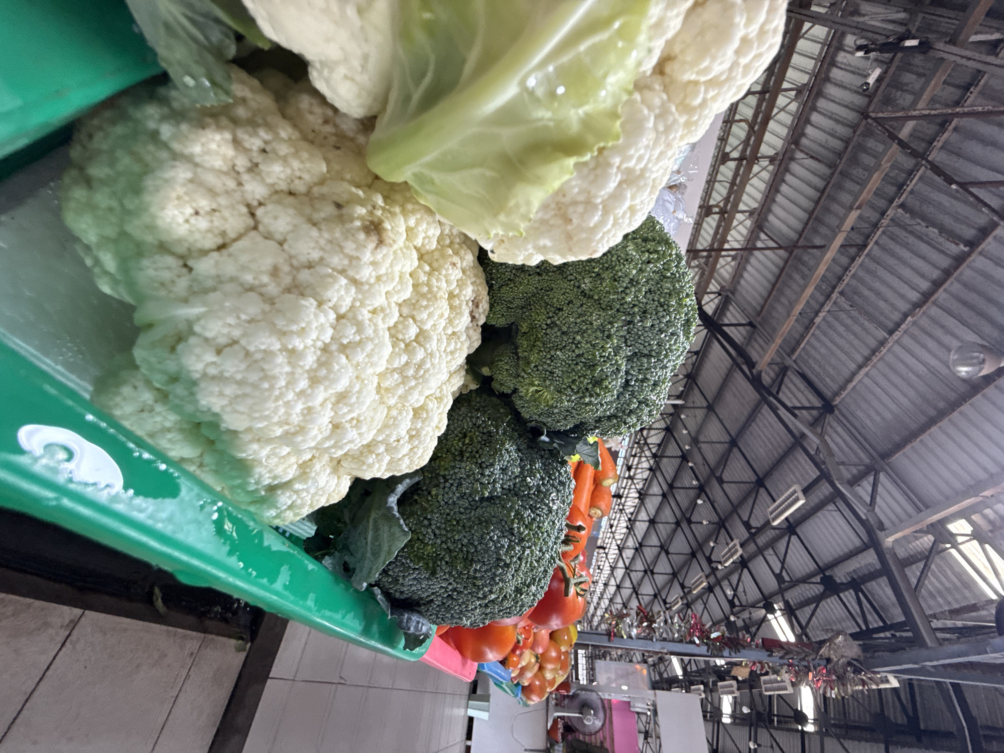 The image displays a close-up, slightly angled view of a selection of fresh vegetables, primarily cauliflower and broccoli, arranged in a green plastic container. The vegetables are the main subjects, appearing fresh and ready for sale or consumption. A green leaf from the cauliflower is visible at the top, overlapping with a head of cauliflower. To the right of the cauliflower, several heads of broccoli are packed tightly together, their florets distinct. Below the broccoli, a cluster of tomatoes and carrots can be seen, adding a splash of color to the arrangement.
The setting appears to be an indoor market or a stall, indicated by the corrugated metal roof structure with exposed beams and lighting fixtures visible in the background. The lighting suggests it might be daytime, though the indoor environment makes it difficult to ascertain natural light conditions or weather. The overall scene is one of fresh produce being displayed in a market setting, likely in Baan Bo Phut, Thailand, as suggested by the provided location context. There are no people clearly visible in the frame, and no text is present. The focus is entirely on the vibrant, textured surfaces of the vegetables and the rustic market architecture.