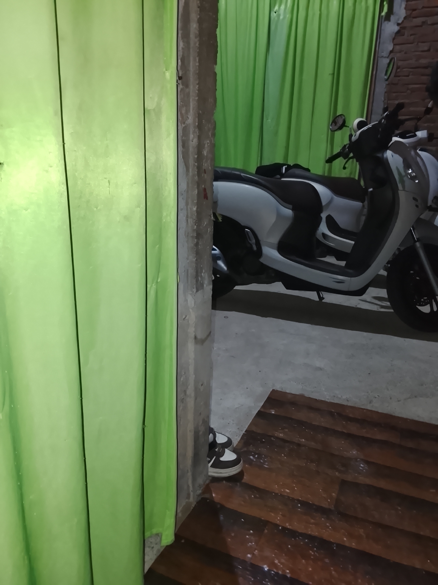 A white and black scooter is parked indoors, partially obscured by a vibrant green curtain. The scooter has a sleek design with a prominent black seat. In the foreground, a pair of black and white sneakers rests on a wooden-patterned floor. The environment appears to be a sheltered space, possibly a garage or a covered outdoor area, with concrete flooring and brickwork visible in the background. The lighting is dim, casting shadows and highlighting the textures of the objects in the scene. The image does not provide explicit details to pinpoint a specific location within Tanjongbabahkrueng, Indonesia, but suggests a residential or commercial setting.